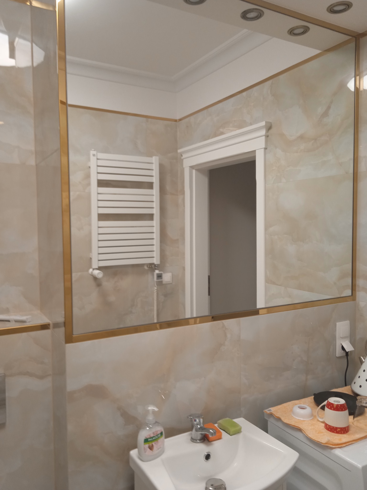 Umywalka z baterią i dozownikiem mydła na tle beżowych płytek w łazience, widoczne lustro z złotą ramą odbijające grzejnik drabinkowy i drzwi.