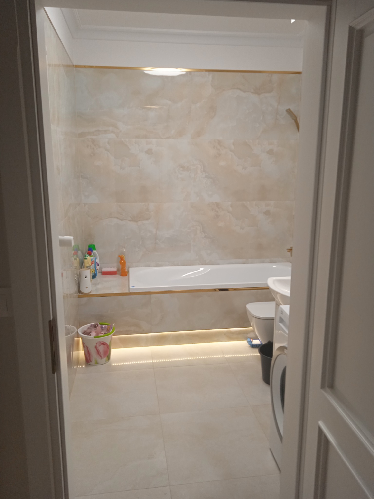 Wykończona łazienka w Bydgoszczy z beżowymi płytkami imitującymi marmur, podświetleniem LED pod wanną i półką, widok zza otwartych drzwi, z widoczną pralką i akcesoriami łazienkowymi.