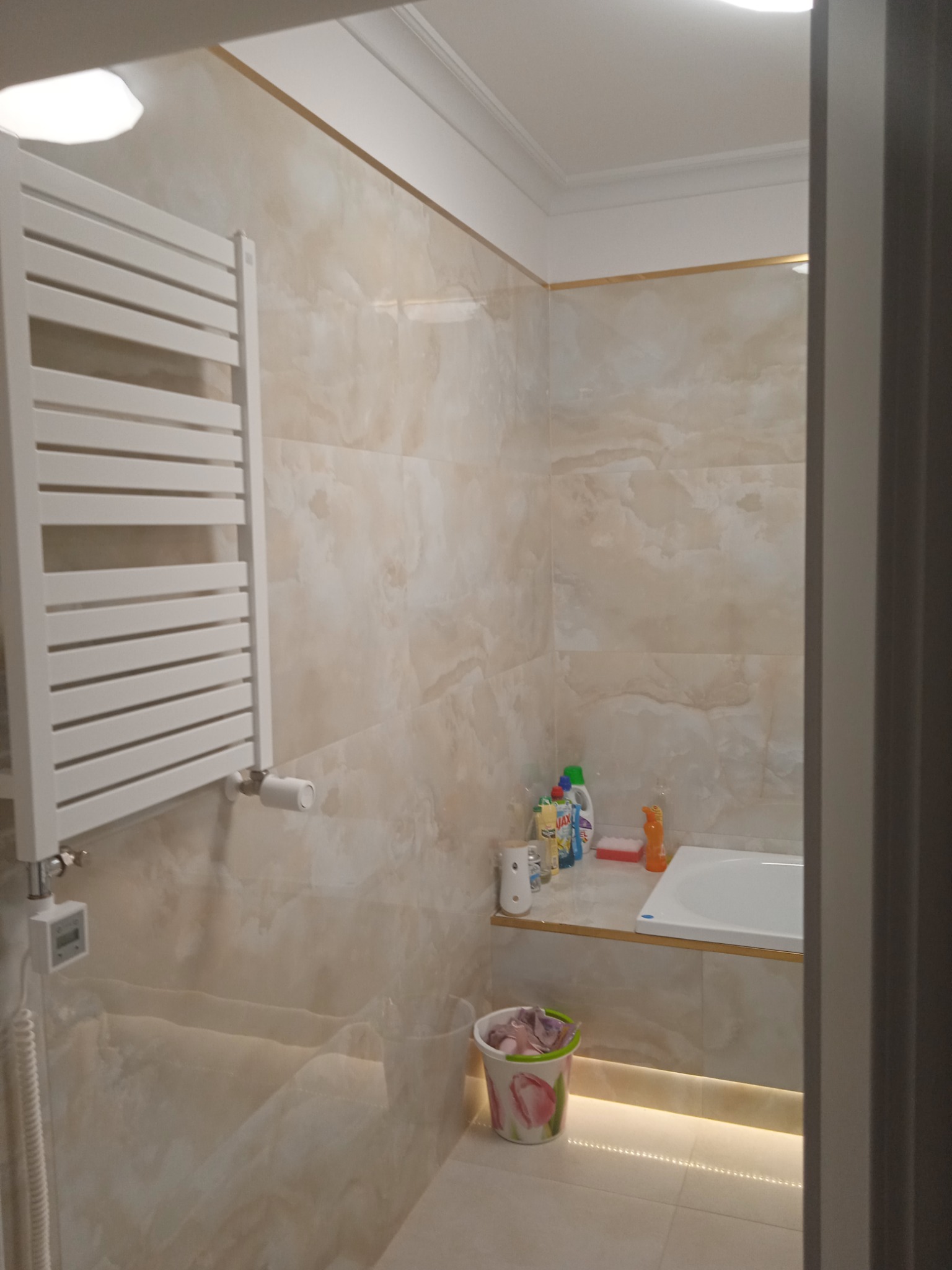 Wykończona łazienka z beżowymi marmurkowymi płytkami, białym grzejnikiem drabinkowym z elektronicznym termostatem, białą wanną i listwą LED pod półką, widok zza framugi drzwi.