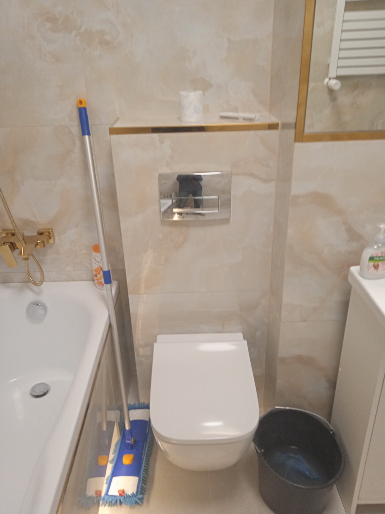 Biała toaleta podwieszana w łazience wykończonej marmurkowymi płytkami, z mopem i wiadrem obok, sugerujące sprzątanie po remoncie.