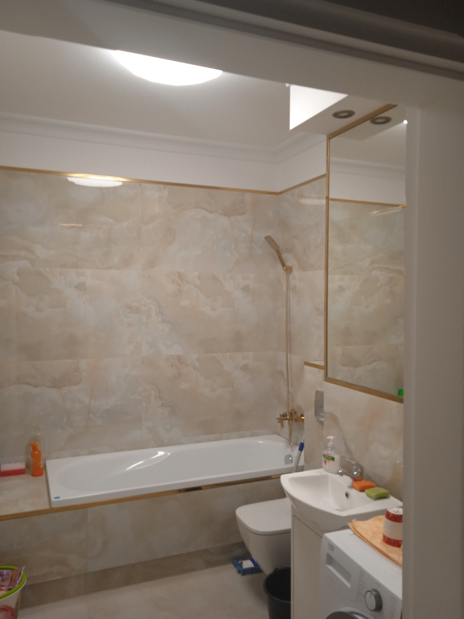 Jasna łazienka z beżowymi płytkami imitującymi marmur, białą wanną, umywalką, toaletą i pralką, wykończona złotymi listwami, oświetlona okrągłą lampą sufitową.