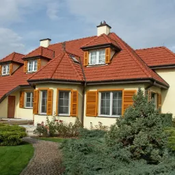 Dwupiętrowy dom z czerwoną dachówką i brązowymi okiennicami, otoczony zielenią i kamienną ścieżką, widziany z poziomu gruntu.