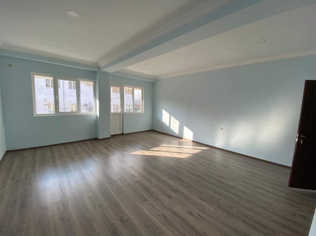 Puste pomieszczenie po remoncie z drewnopodobnymi panelami podłogowymi, świeżo pomalowanymi błękitnymi ścianami, białym sufitem z dekoracyjną listwą i trzema oknami w białych ramach.