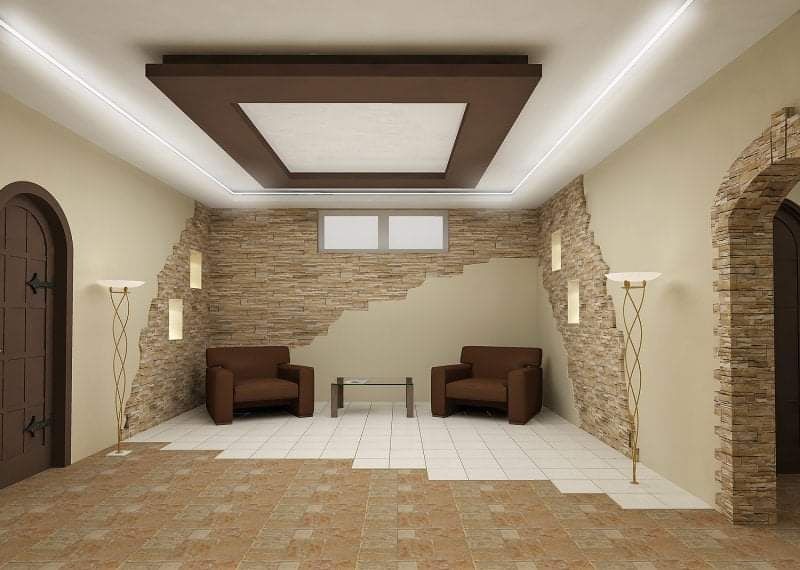 Wnętrze pokoju z częściowo wykończoną ścianą z kamienia dekoracyjnego, brązowymi fotelami i podłogą z częściowo ułożonymi płytkami w dwóch kolorach, oświetlenie sufitowe z brązową ramą.