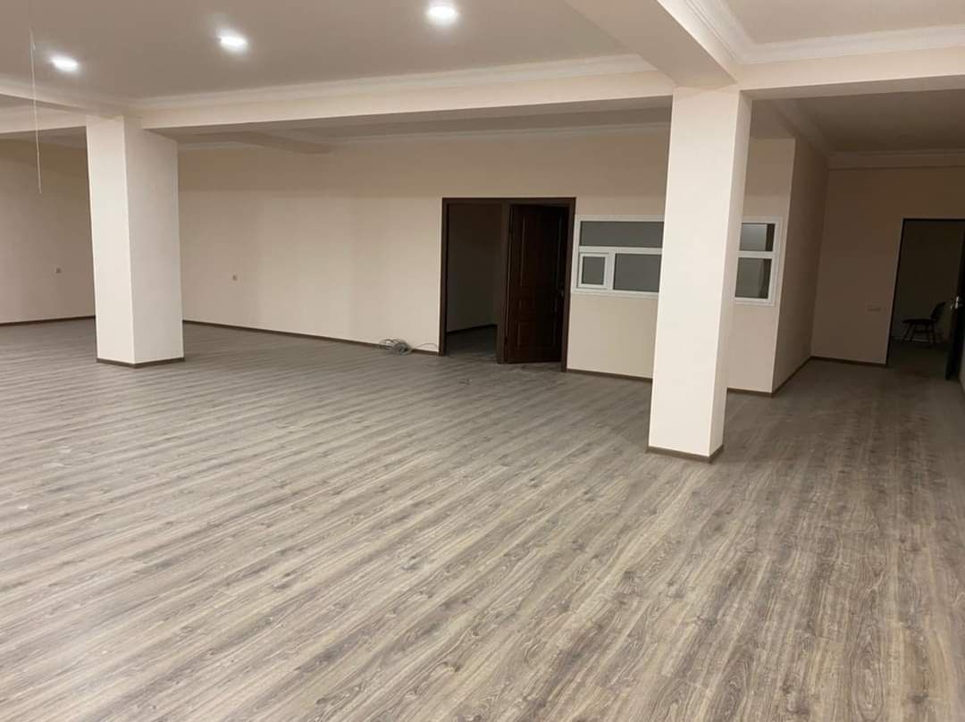 Jasne wnętrze z szarymi panelami podłogowymi ułożonymi wzdłużnie, białymi kolumnami i beżowymi ścianami, widoczne drzwi i okno.