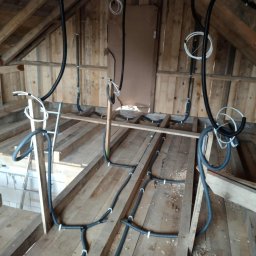 PD-SYSTEM PATRYK DOLECKI - Instalacja przewodów w budynku w trakcie budowy. Widoczne kable prowadzone po drewnianej konstrukcji poddasza, częściowo przymocowane uchwytami. Surowy stan wykończenia.