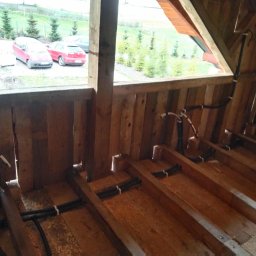 PD-SYSTEM PATRYK DOLECKI - Instalacja elektryczna w drewnianej konstrukcji poddasza, widoczne kable przymocowane do belek, okno z widokiem na zewnątrz. Prace instalacyjne w trakcie realizacji.