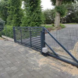 Przesuwna brama wjazdowa z automatyką, widoczny napęd, szara kolorystyka, kostka brukowa na podjeździe, w tle zieleń drzew i ogrodzenie panelowe.