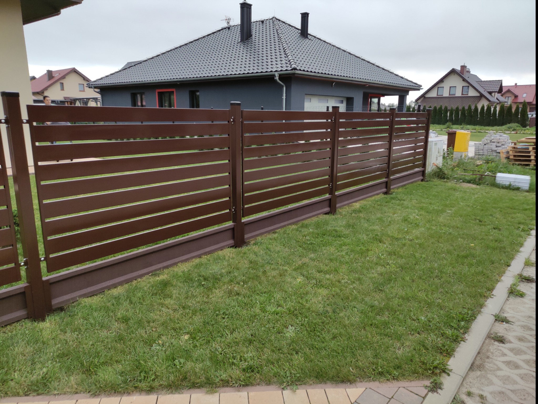Nowoczesne, poziome ogrodzenie panelowe w kolorze czekoladowego brązu, oddzielające posesję z trawnikiem od ulicy, widoczne domy w tle.
