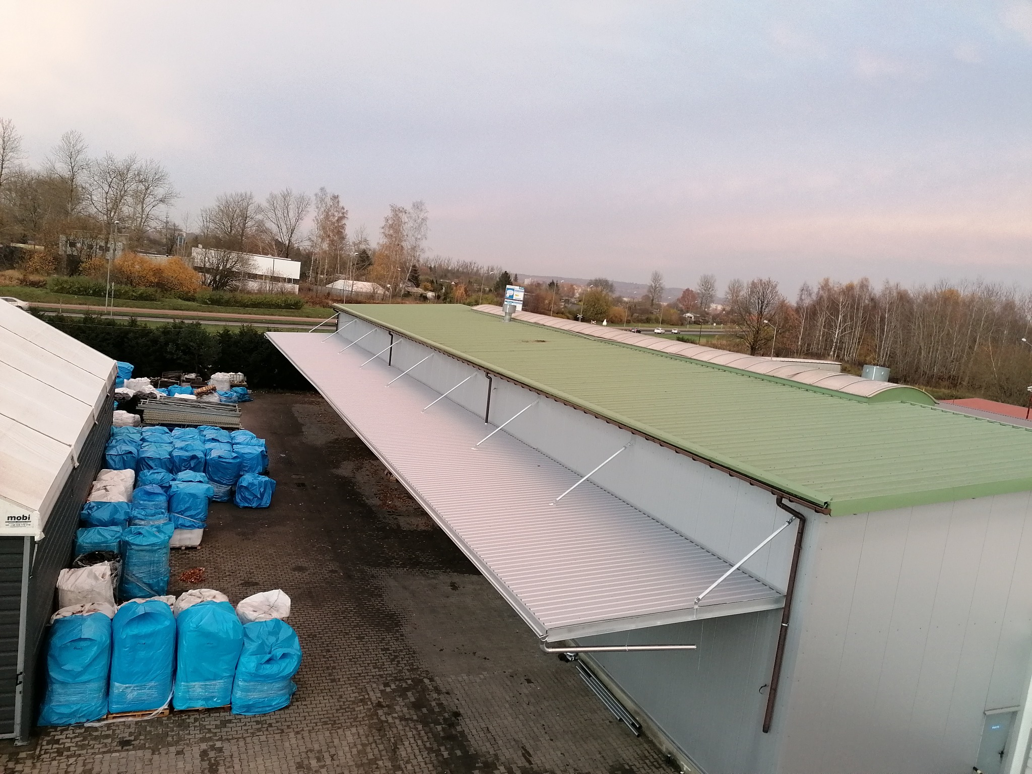 Widok z góry na nowo wybudowaną halę z zielonym dachem i wysuwanym zadaszeniem, obok sterty niebieskich worków na paletach.