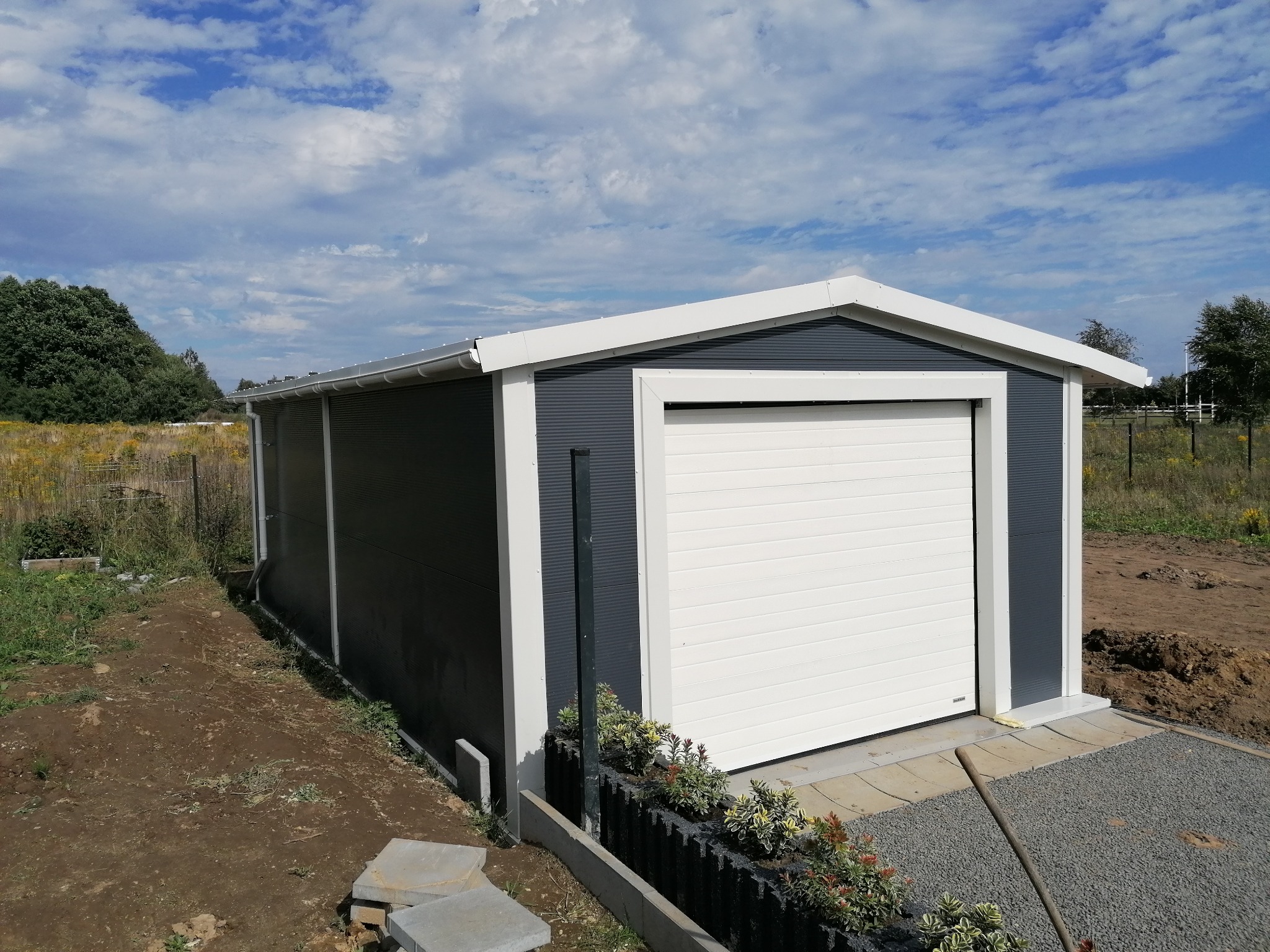 Gotowa hala garażowa z szarą blachą trapezową i białą bramą segmentową, świeżo po montażu, z widocznymi elementami zagospodarowania terenu wokół.