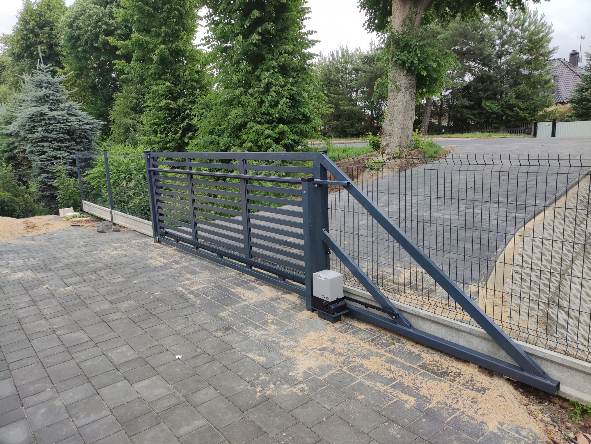 Przesuwna brama wjazdowa z automatyką, widoczny napęd, szara kolorystyka, kostka brukowa na podjeździe, w tle zieleń drzew i ogrodzenie panelowe.