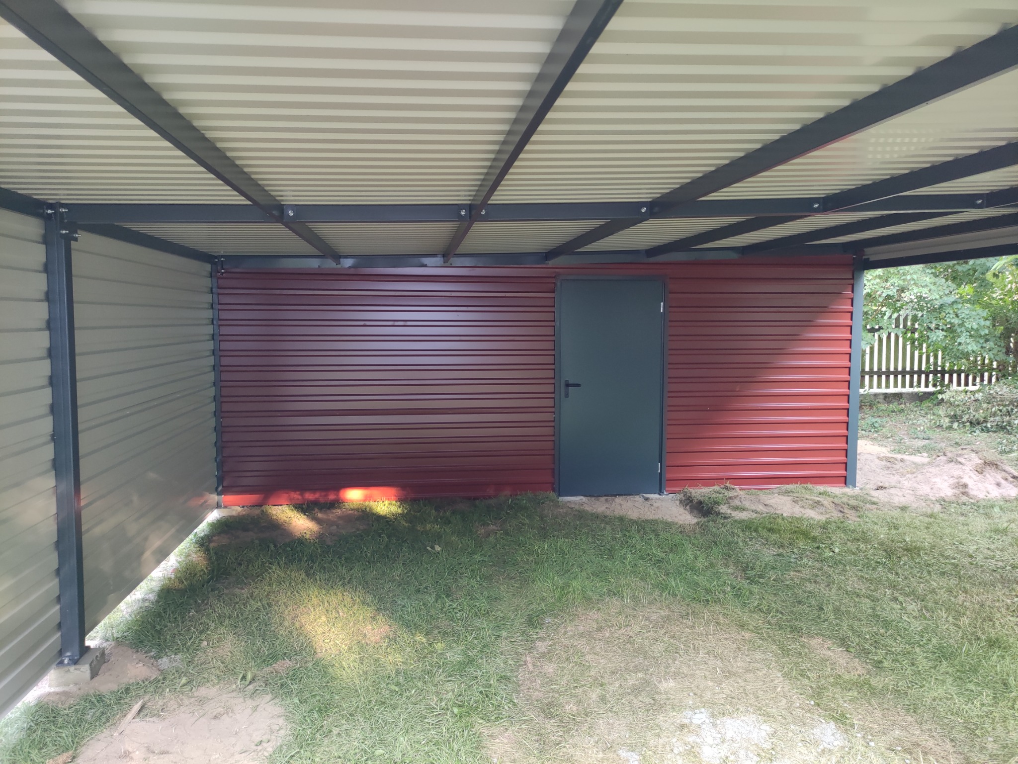 Zadaszona konstrukcja z blachy trapezowej w kolorze bordowym i szarym, z ciemnoszarymi słupami nośnymi i drzwiami wejściowymi, na zewnątrz budynku.