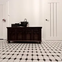 Wzorzysta podłoga z białych i czarnych płytek ułożonych w geometryczny wzór, na której stoi ciemnobrązowa, zdobiona skrzynia na tle dwóch białych drzwi.