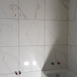 Mateusz Barański 3M BAU - Ściany łazienki wyłożone białymi płytkami imitującymi marmur, widoczne instalacje wodne i elektryczne przed montażem armatury, surowy narożnik ściany z widocznym betonem.