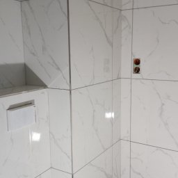 Mateusz Barański 3M BAU - Ściana łazienki wyłożona białymi płytkami imitującymi marmur z widocznymi przyłączami wodno-kanalizacyjnymi i zamontowaną spłuczką podtynkową.