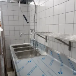 Profesjonalna, trzydzielna zlewozmywarka ze stali nierdzewnej z baterią przemysłową i półką na tle białych płytek.