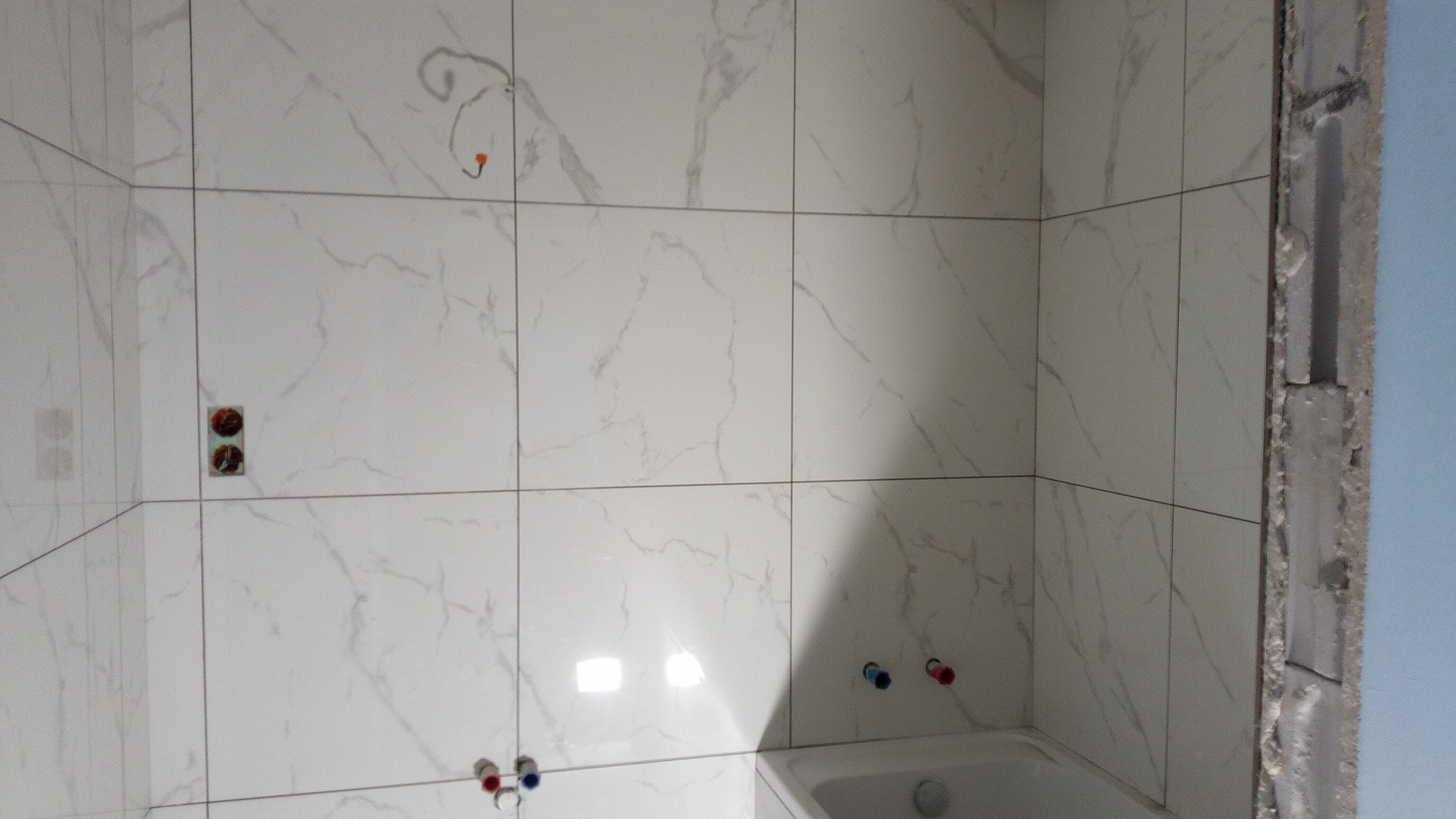 Ściany łazienki wyłożone białymi płytkami imitującymi marmur, widoczne instalacje wodne i elektryczne przed montażem armatury, surowy narożnik ściany z widocznym betonem.