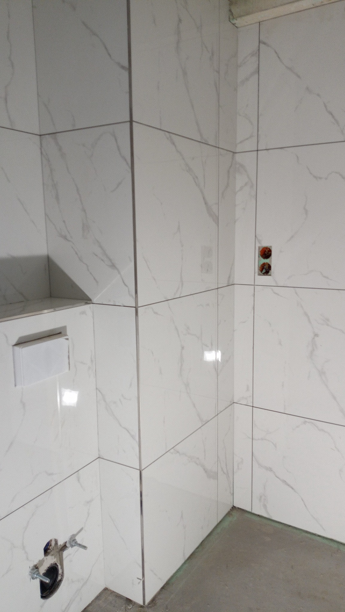 Ściana łazienki wyłożona białymi płytkami imitującymi marmur z widocznymi przyłączami wodno-kanalizacyjnymi i zamontowaną spłuczką podtynkową.