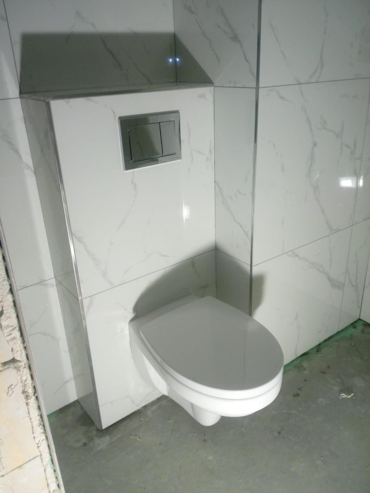 Wisząca toaleta z ukrytym stelażem, obudowana białymi płytkami imitującymi marmur, z chromowanym przyciskiem spłukującym.