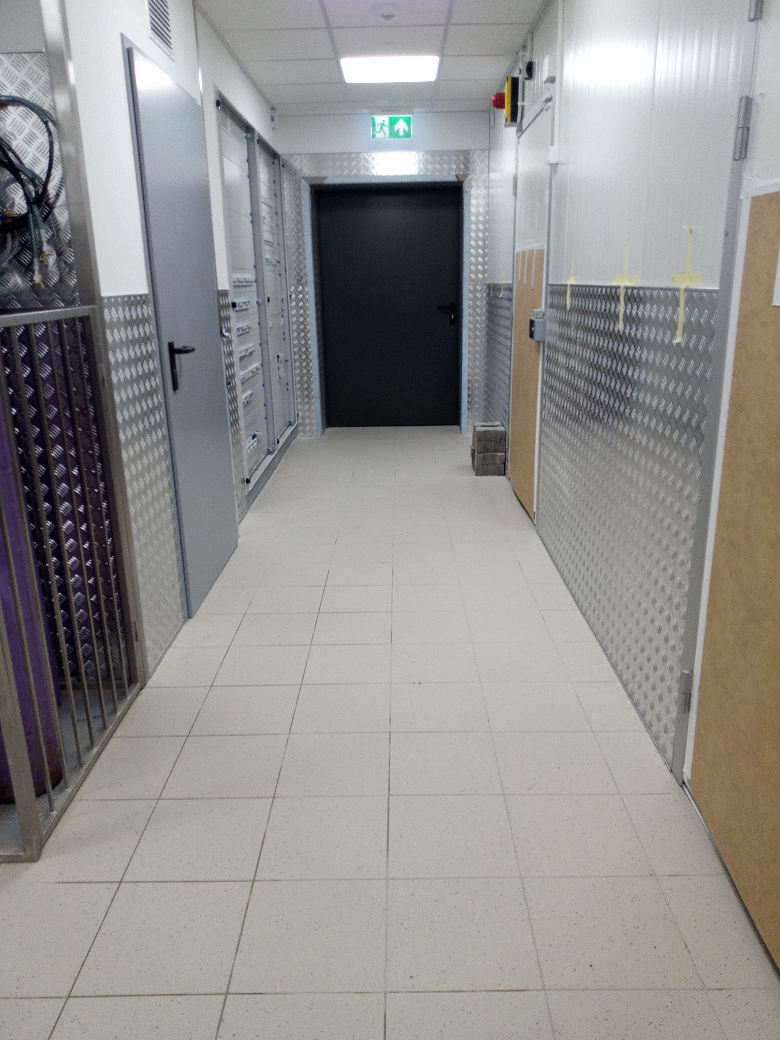 Korytarz w budynku przemysłowym z białymi płytkami na podłodze, ścianami wyłożonymi blachą ryflowaną i szarymi drzwiami oraz czarnymi drzwiami ewakuacyjnymi na końcu.