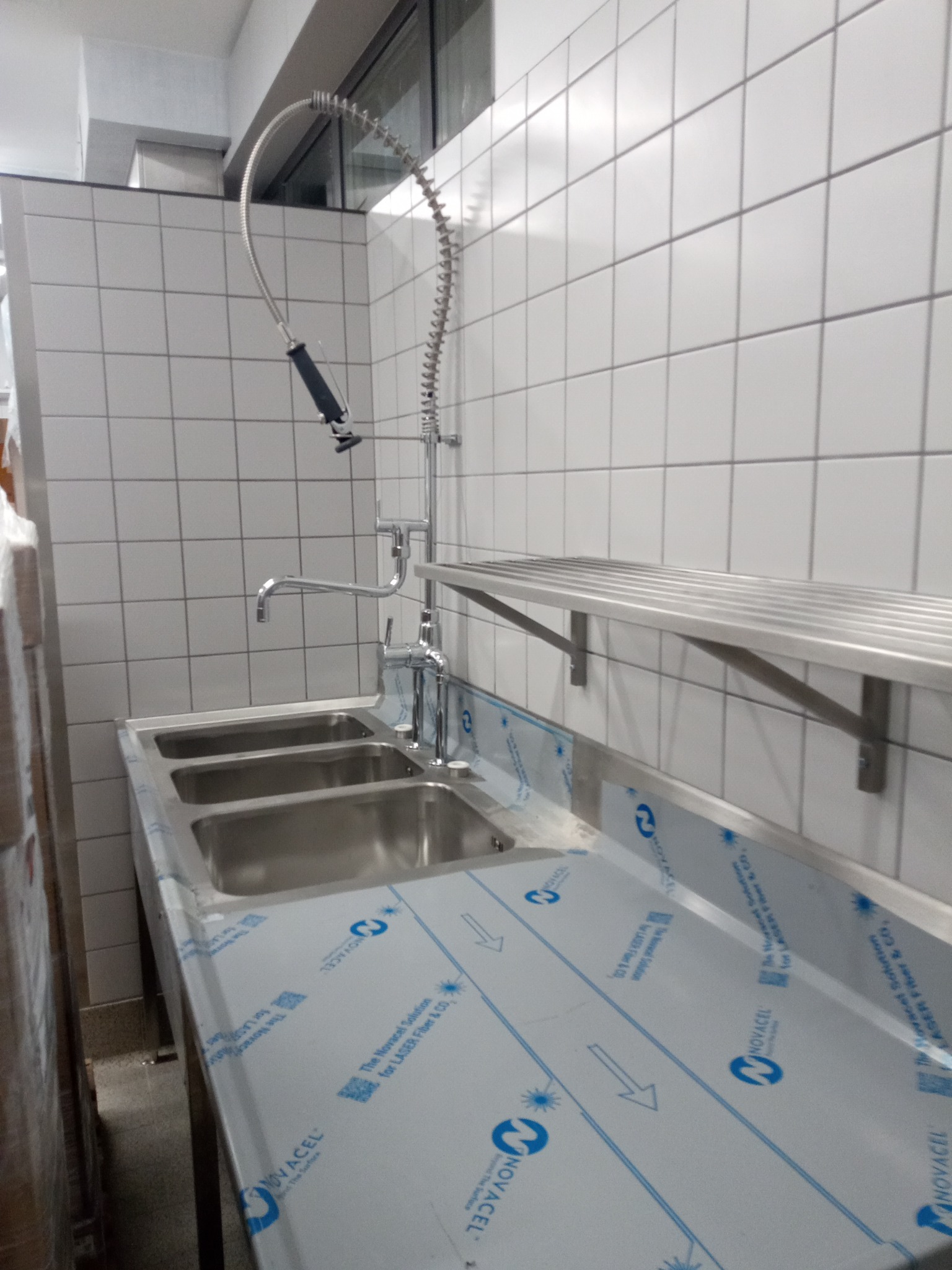 Profesjonalna, trzydzielna zlewozmywarka ze stali nierdzewnej z baterią przemysłową i półką na tle białych płytek.