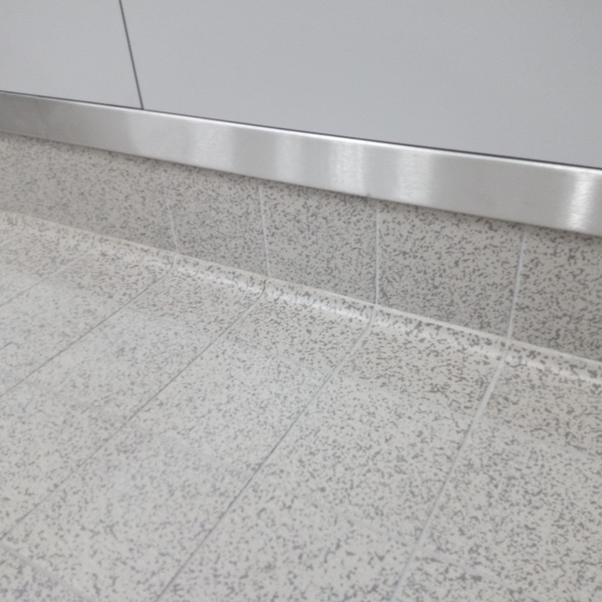 Jasnoszara, nakrapiana podłoga z płytek w Legnicy, wykończona metalową listwą przypodłogową, przylegająca do gładkiej, jasnej ściany.