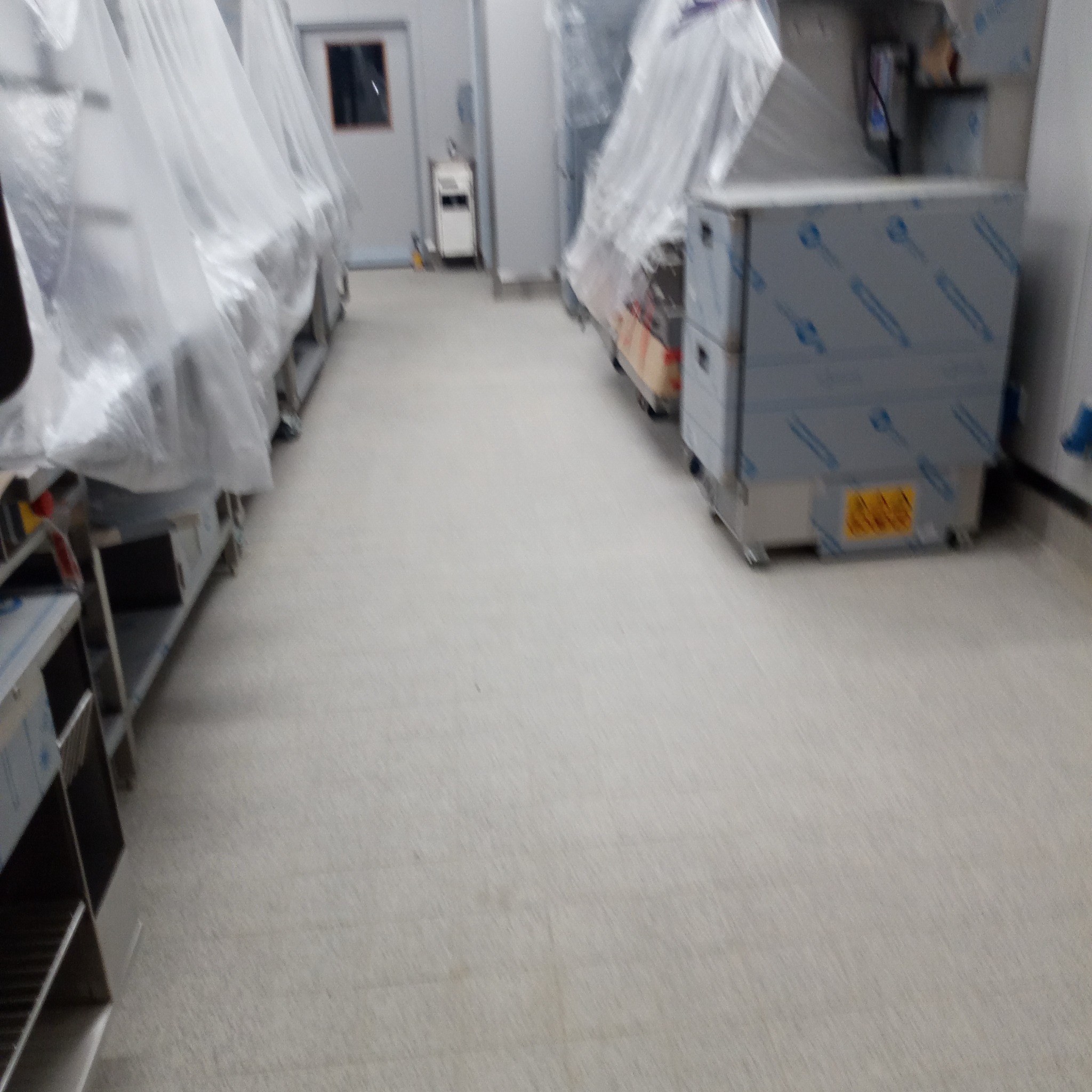 Wnętrze hali produkcyjnej z nowymi urządzeniami przykrytymi folią ochronną, widoczna podłoga z płytek i białe ściany.