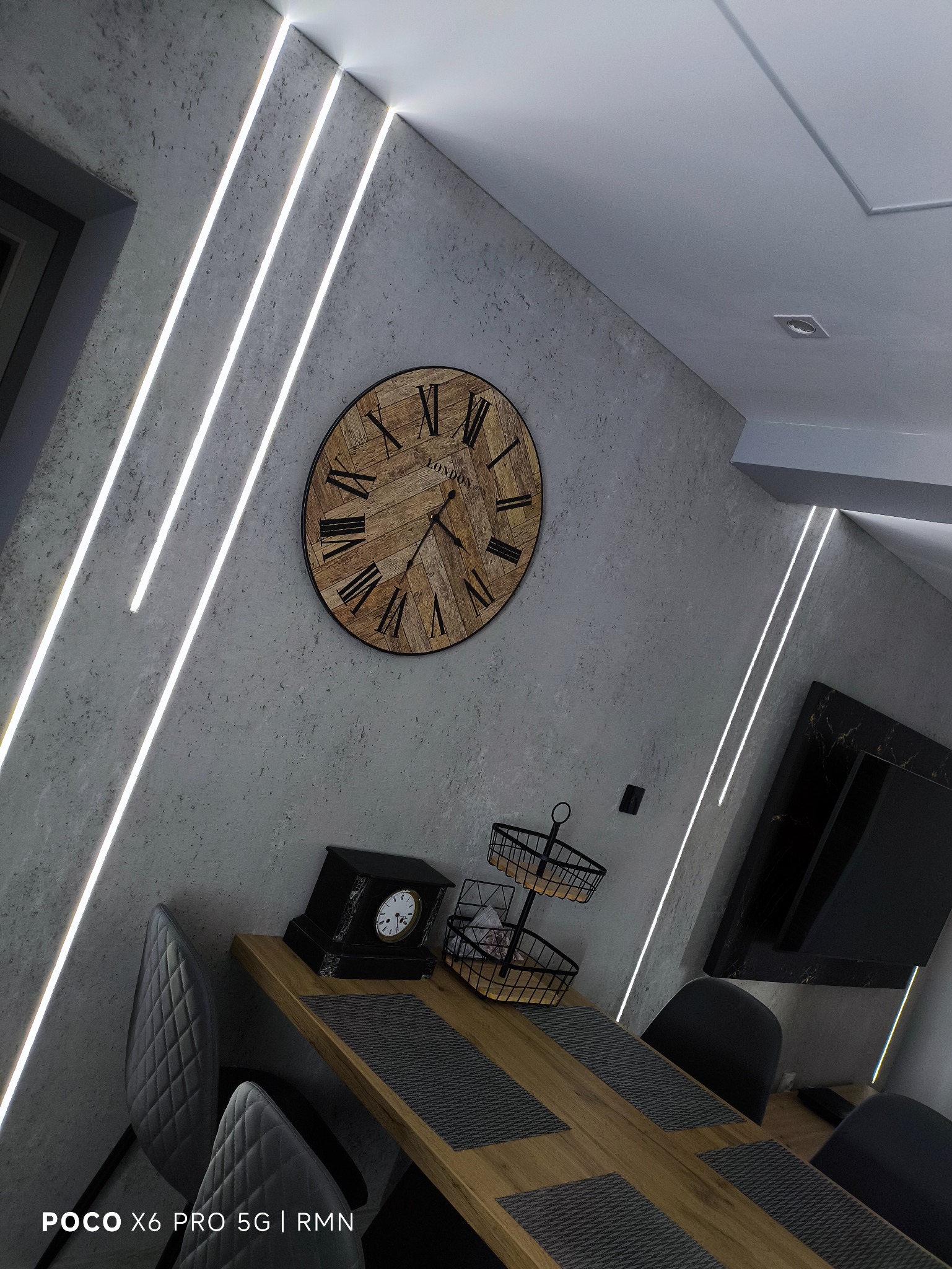 Betonowa ściana z dekoracyjnym zegarem i oświetleniem LED, stół z szarymi podkładkami i krzesła. Nowoczesne wykończenie wnętrza z dbałością o detale.