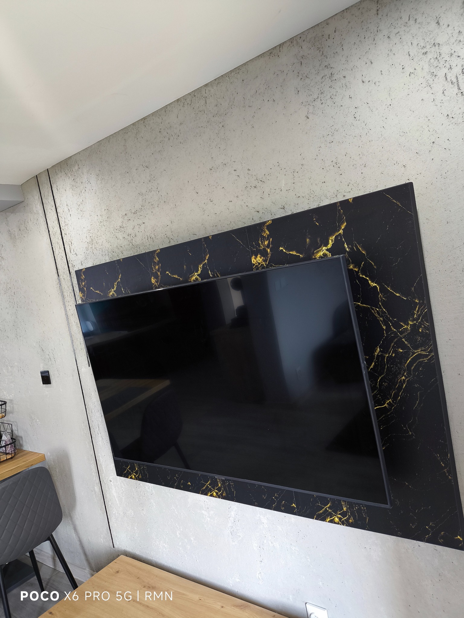 Telewizor w czarnej, marmurowej obudowie z złotymi żyłkami, zamontowany na szarej ścianie o surowym wykończeniu. Widoczny fragment blatu i krzesła.