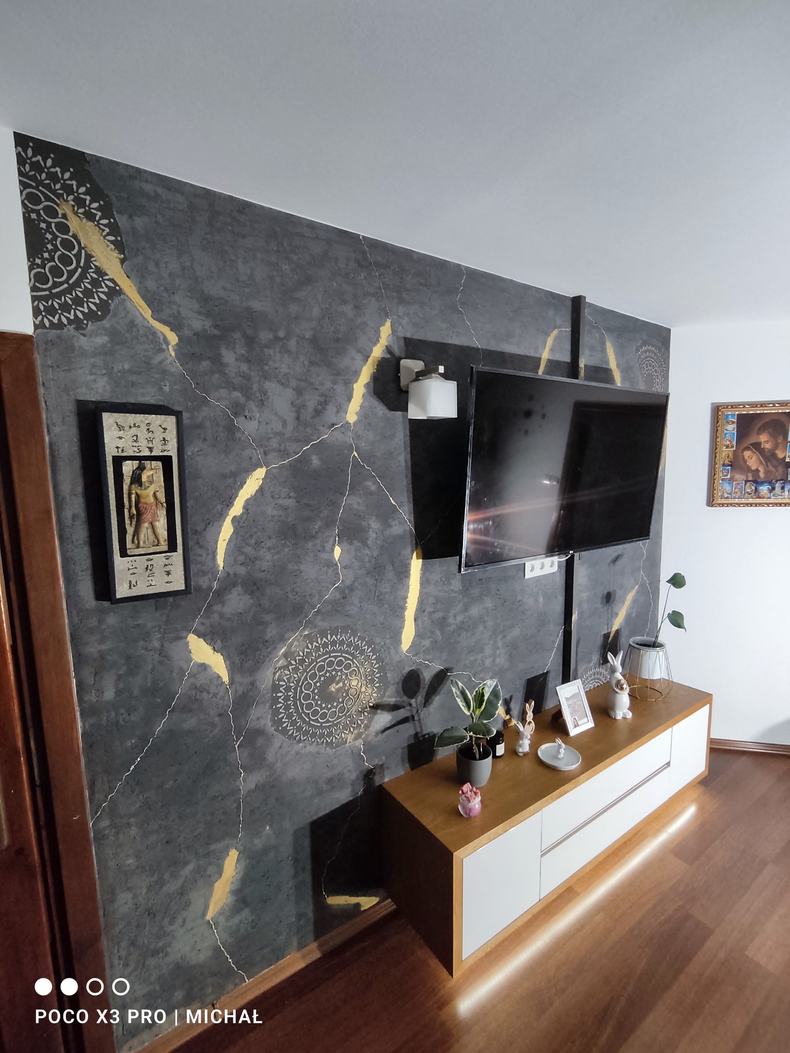 Ściana w salonie z dekoracyjną, szarą fakturą, złotymi żyłkami i ornamentami. Na ścianie wisi telewizor i obrazek w ramce. Pod ścianą szafka RTV z dekoracjami.