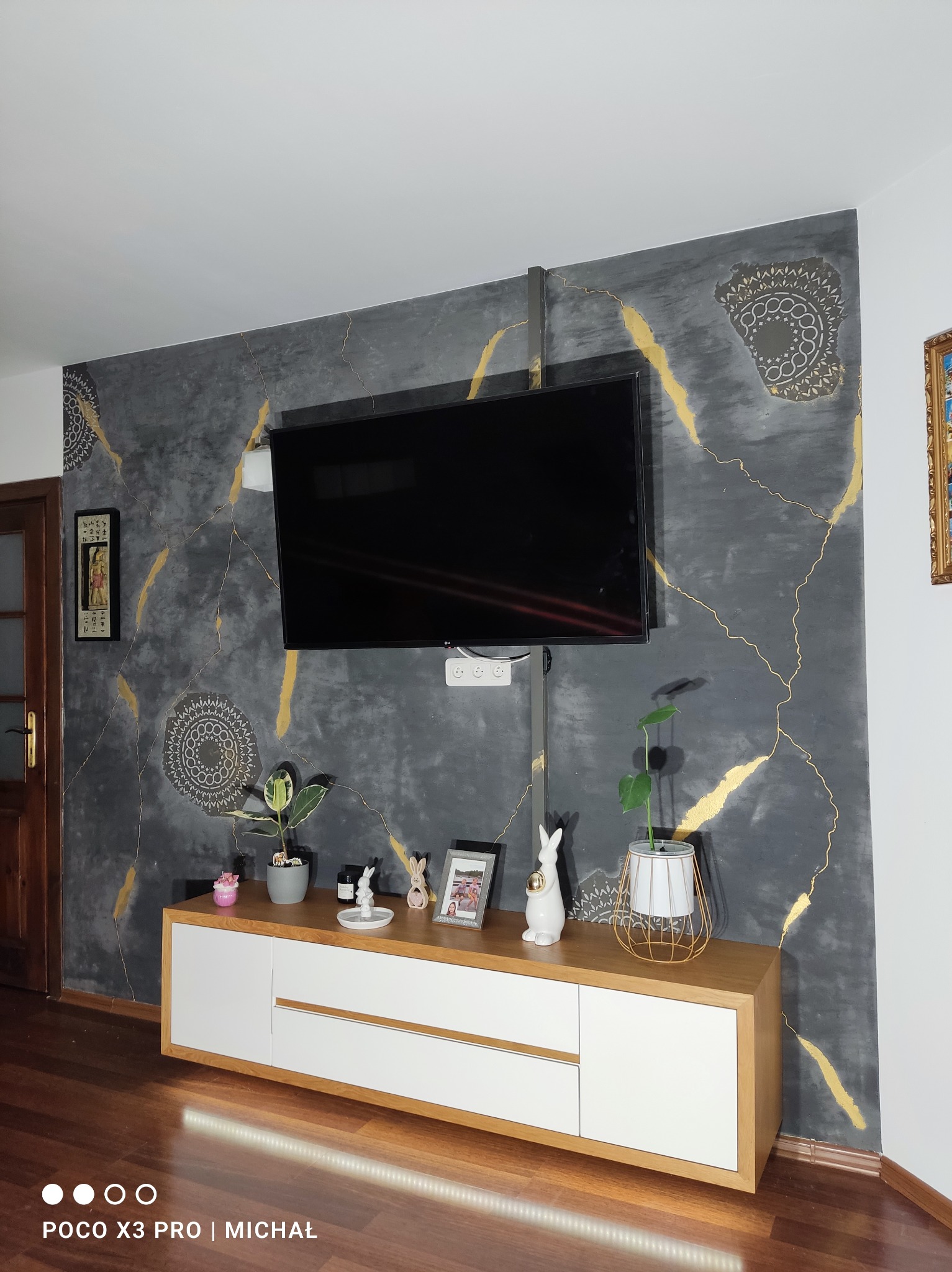 Ściana w salonie z efektem betonu i złotymi żyłkami, dekoracyjne mandale, szafka RTV z roślinami. Nowoczesny design wnętrza.