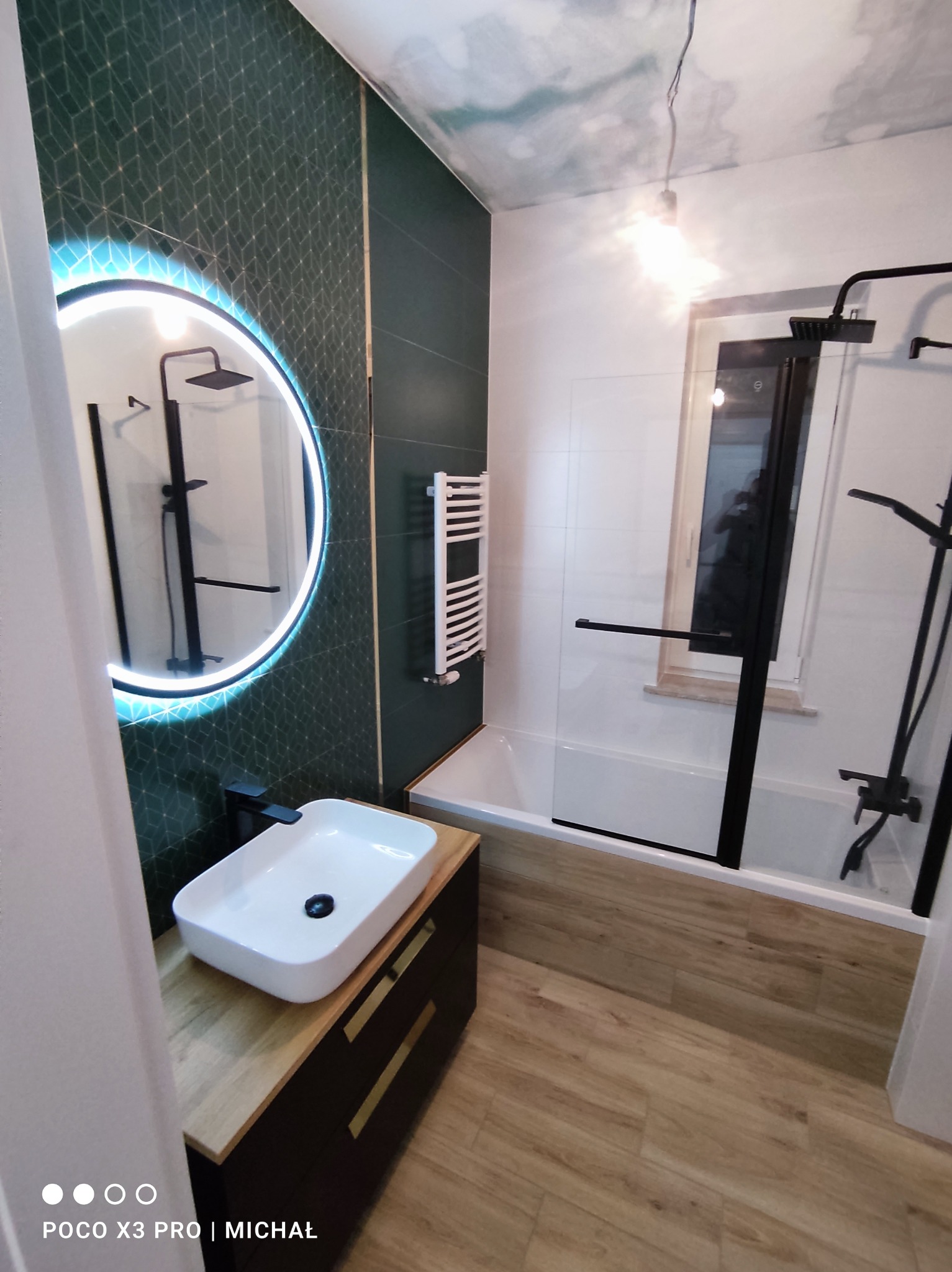 Nowoczesna łazienka po remoncie: zielone płytki z geometrycznym wzorem, okrągłe lustro z podświetleniem LED, biała umywalka nablatowa i kabina prysznicowa.