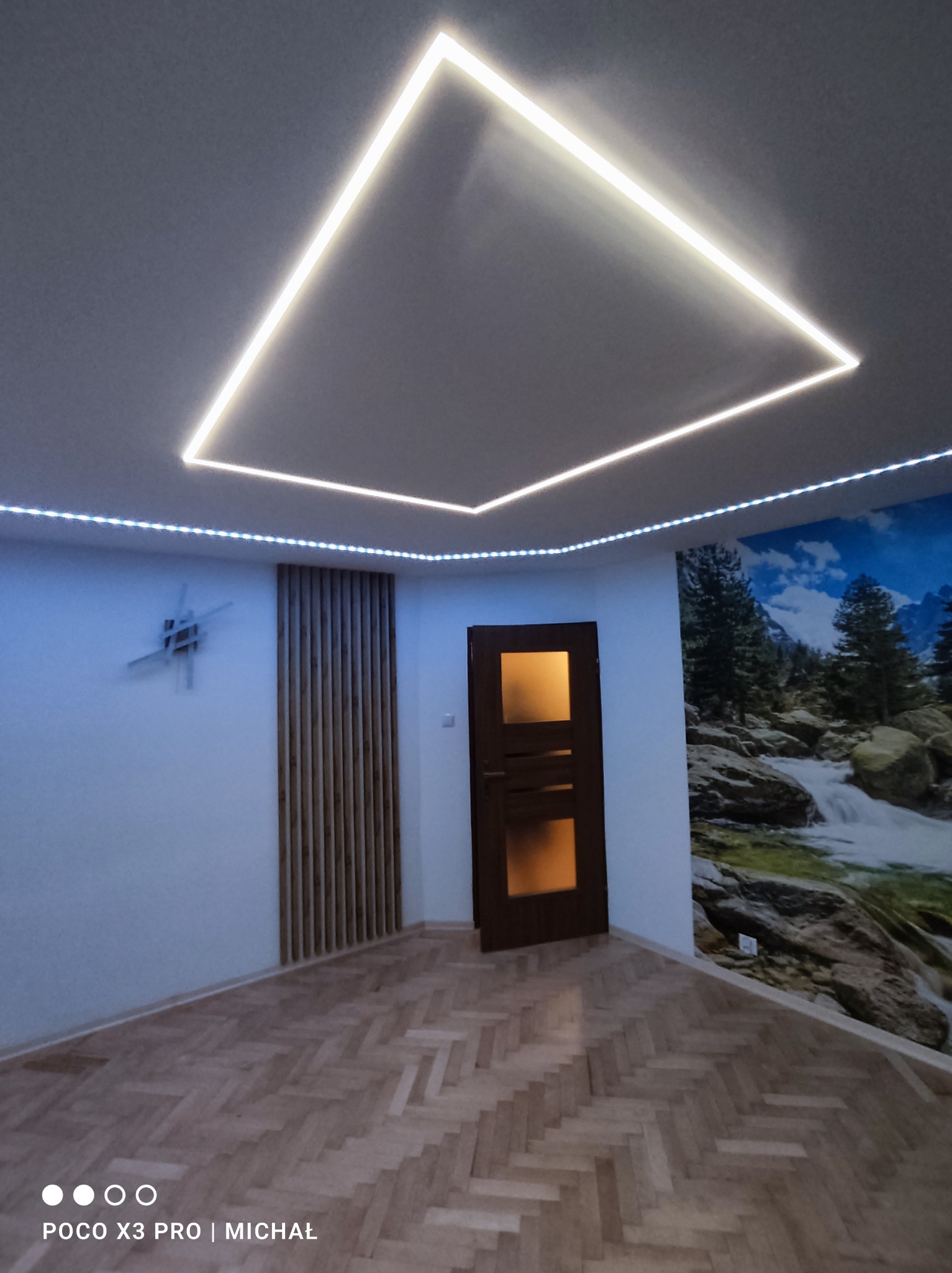 Pokój z parkietem w jodełkę, podświetlonym sufitem z ledowym kwadratem i paskiem, drewnianą ścianką działową oraz fototapetą z motywem górskiego wodospadu.