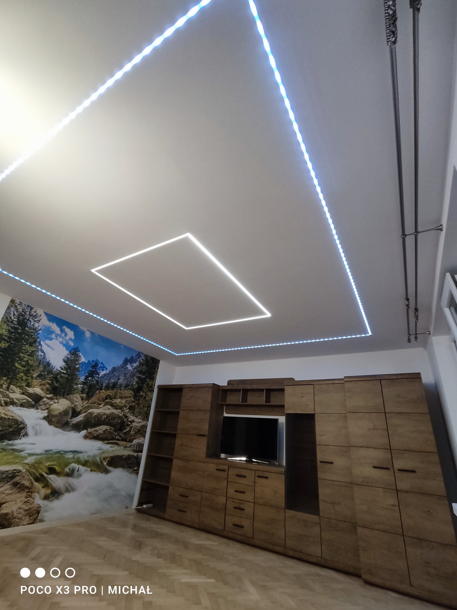 Nowoczesne oświetlenie LED sufitowe w kształcie kwadratu i prostokąta, z tapetą z motywem wodospadu i drewnianą zabudową meblową w tle. Nietypowe rozwiązanie oświetleniowe.