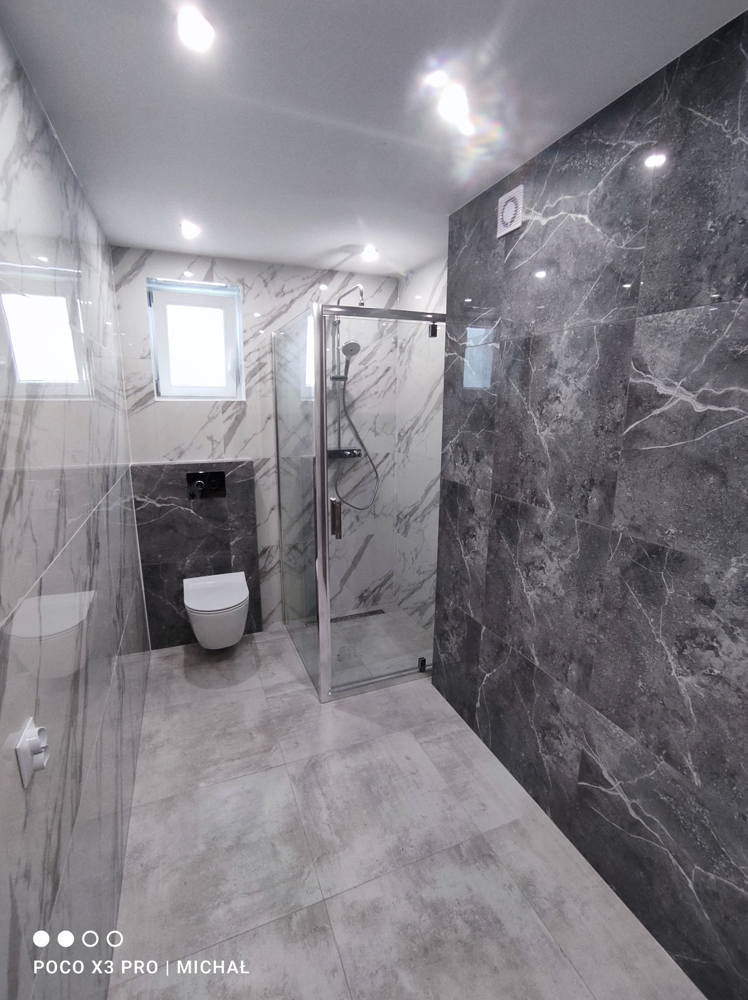 Nowoczesna łazienka po remoncie: szare płytki imitujące marmur na ścianach i podłodze, kabina prysznicowa ze szkła, podwieszana toaleta. Minimalistyczny design.