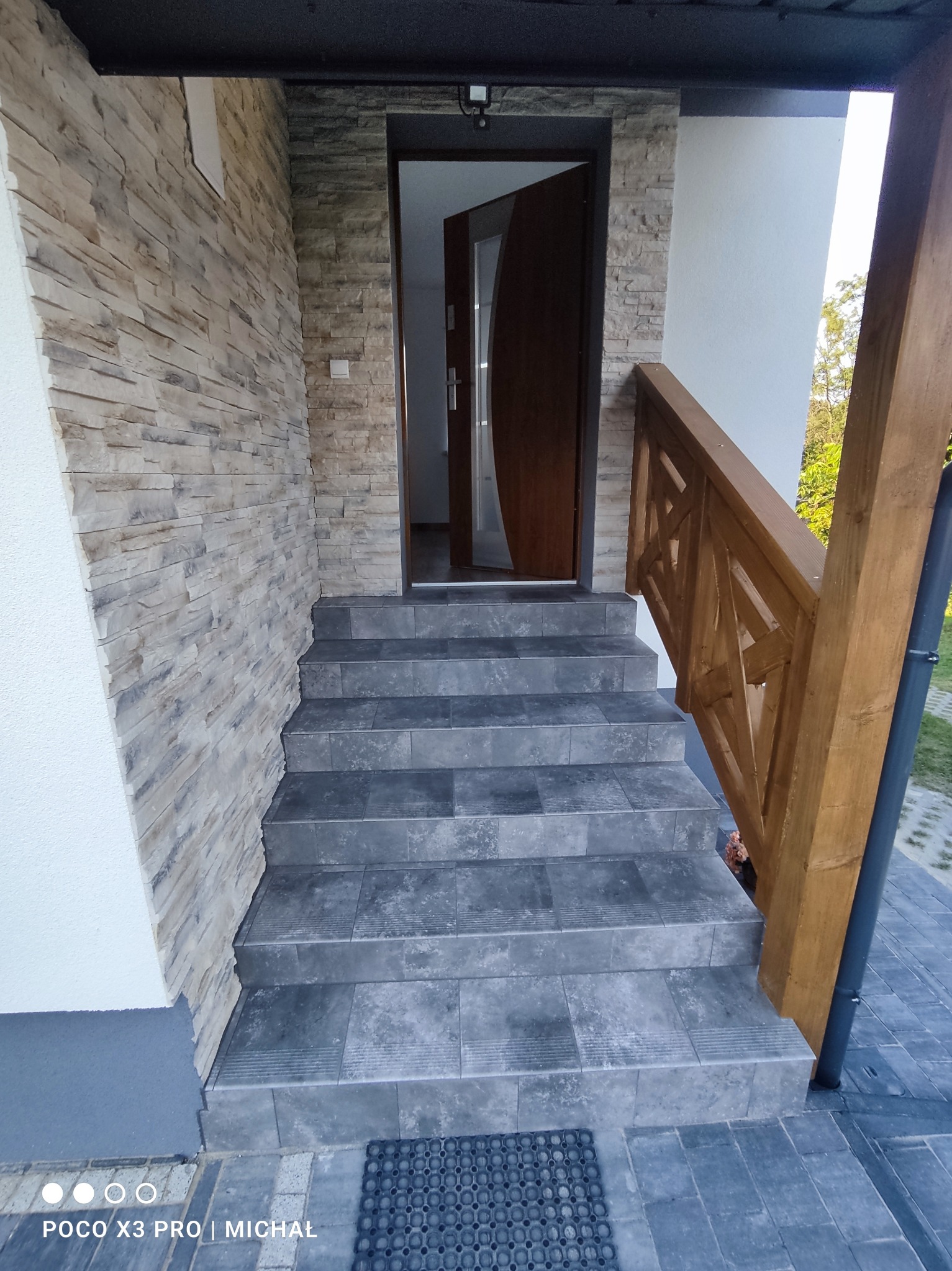 Wykończone schody wejściowe z szarych płytek, drewniana balustrada z geometrycznym wzorem, elewacja z kamienia dekoracyjnego. Widok na otwarte drzwi wejściowe.