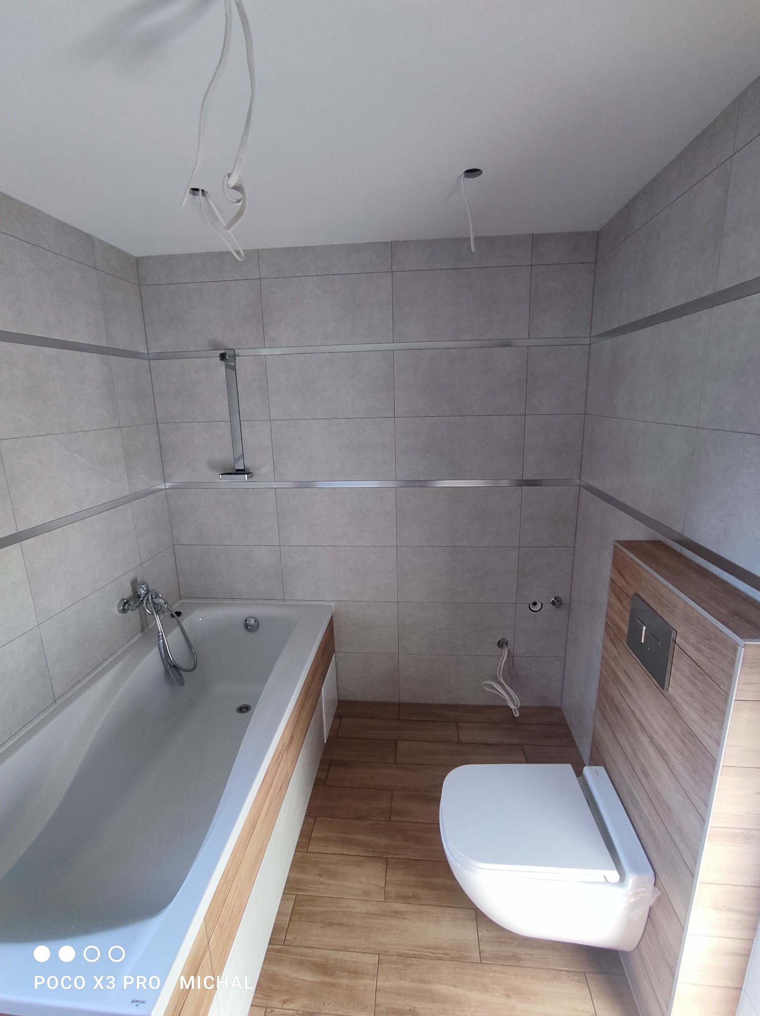 Wykończona łazienka z beżowymi płytkami, drewnopodobną podłogą i wiszącą toaletą. Widoczne niedokończone podłączenia elektryczne.