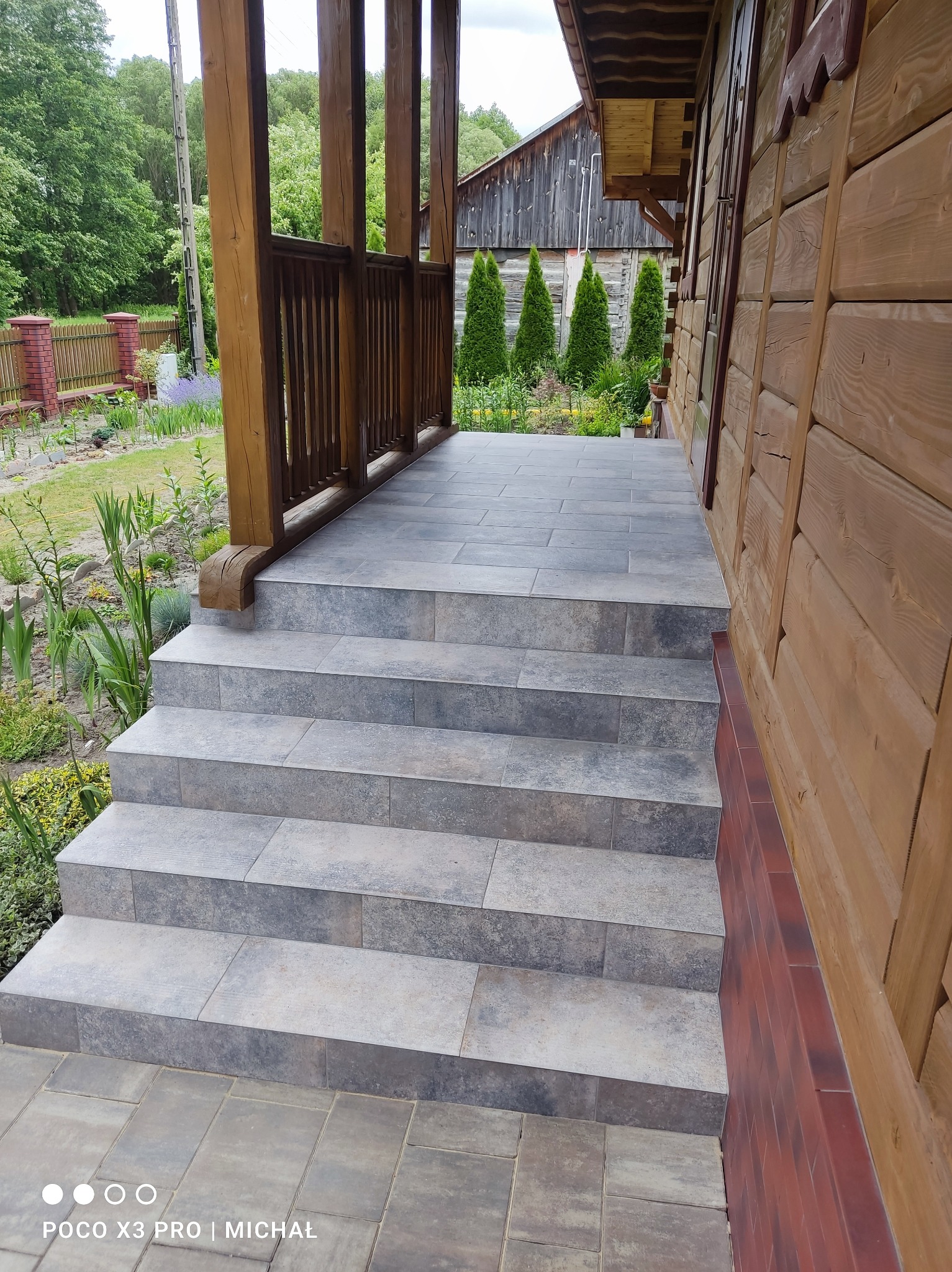 Wyłożone płytkami schody wejściowe do drewnianego domu z balustradą, otoczone zielenią. Widoczne płytki w odcieniach szarości i brązu, dopasowane do elewacji.