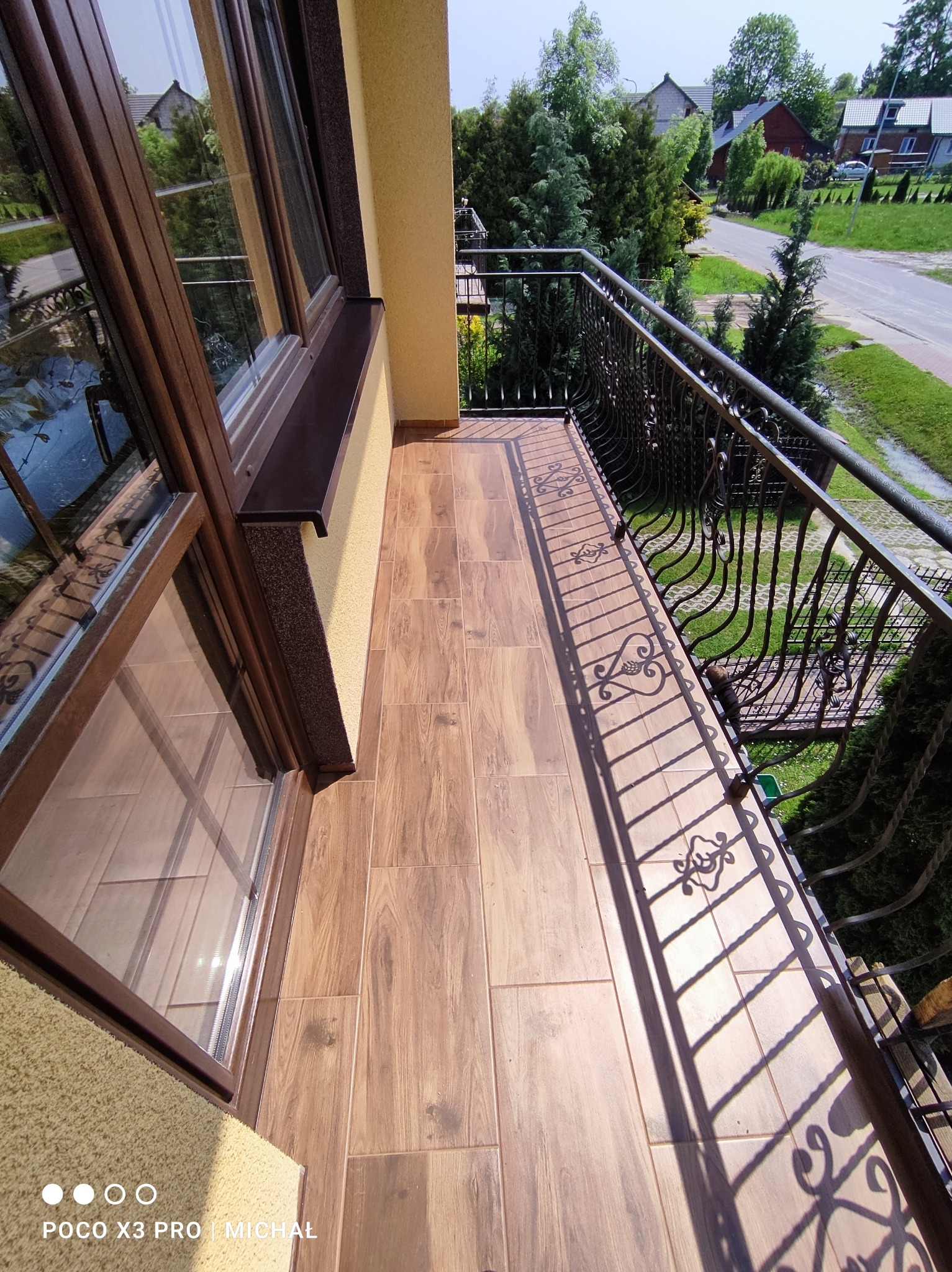 Balkon z brązowymi płytkami imitującymi drewno, ozdobna metalowa balustrada rzucająca cień na podłogę. Widok na zieleń i domy w tle, okno z brązową ramą.