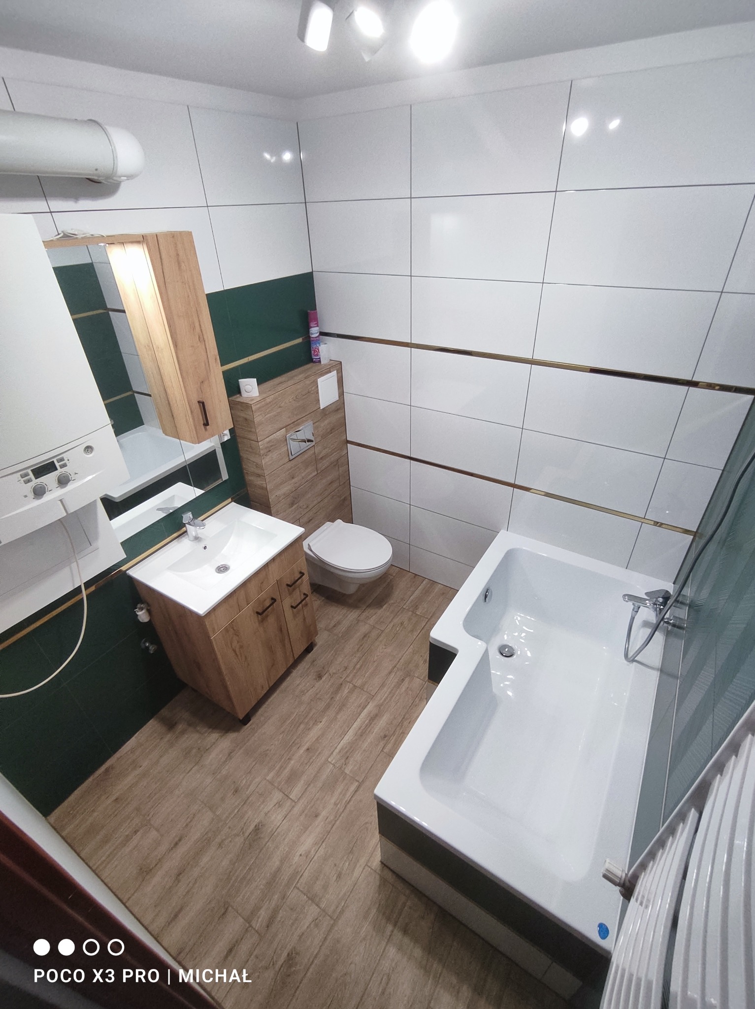 Wykończona łazienka w Chyżem z wanną, umywalką z szafką w odcieniach drewna i toaletą. Białe płytki z poziomymi złotymi paskami i zielony pas na ścianach.