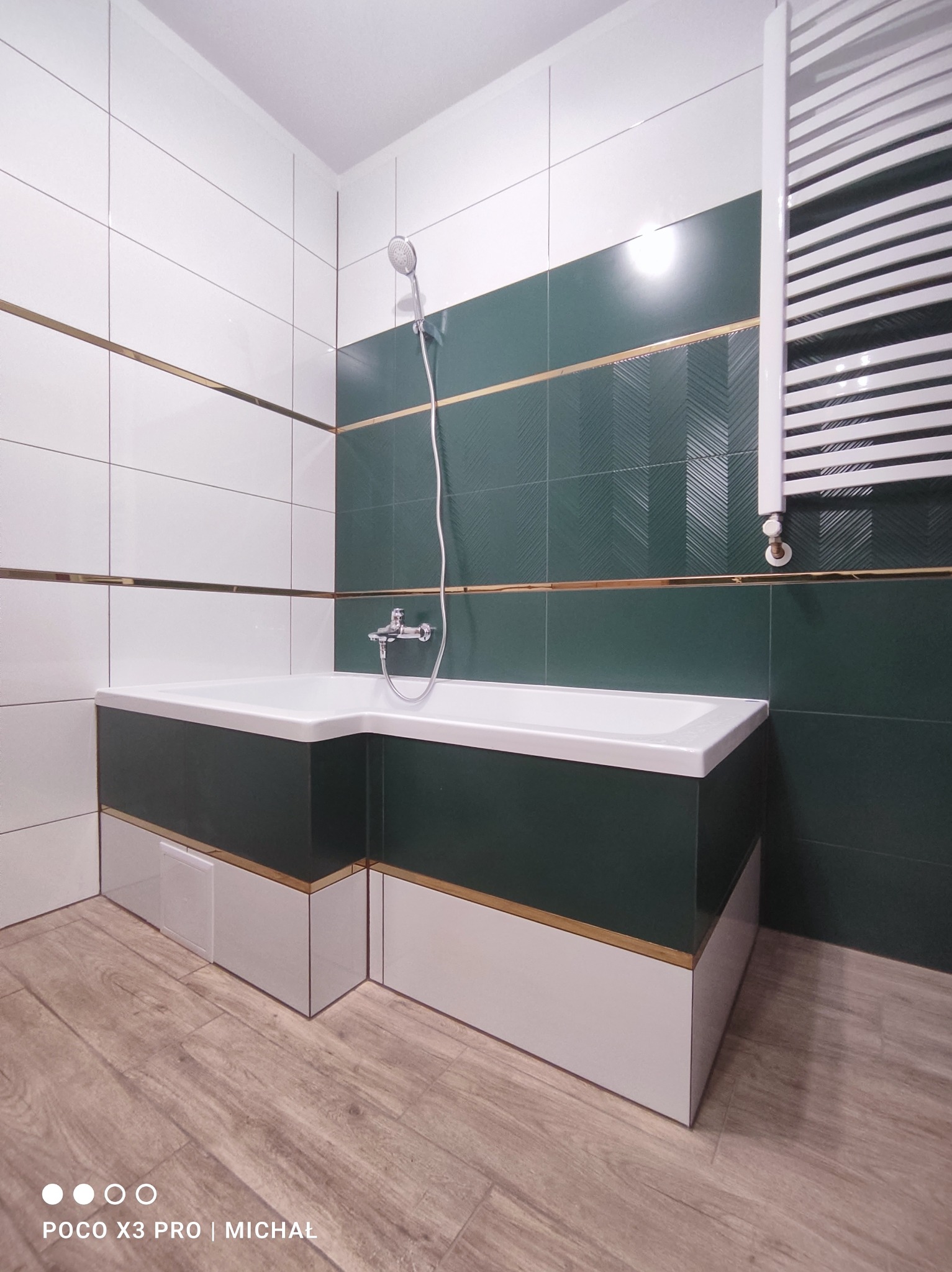 Nowoczesna łazienka z wanną obudowaną zielono-białymi płytkami, akcenty złota, białe płytki na ścianach i drewnopodobna podłoga. Grzejnik drabinkowy.