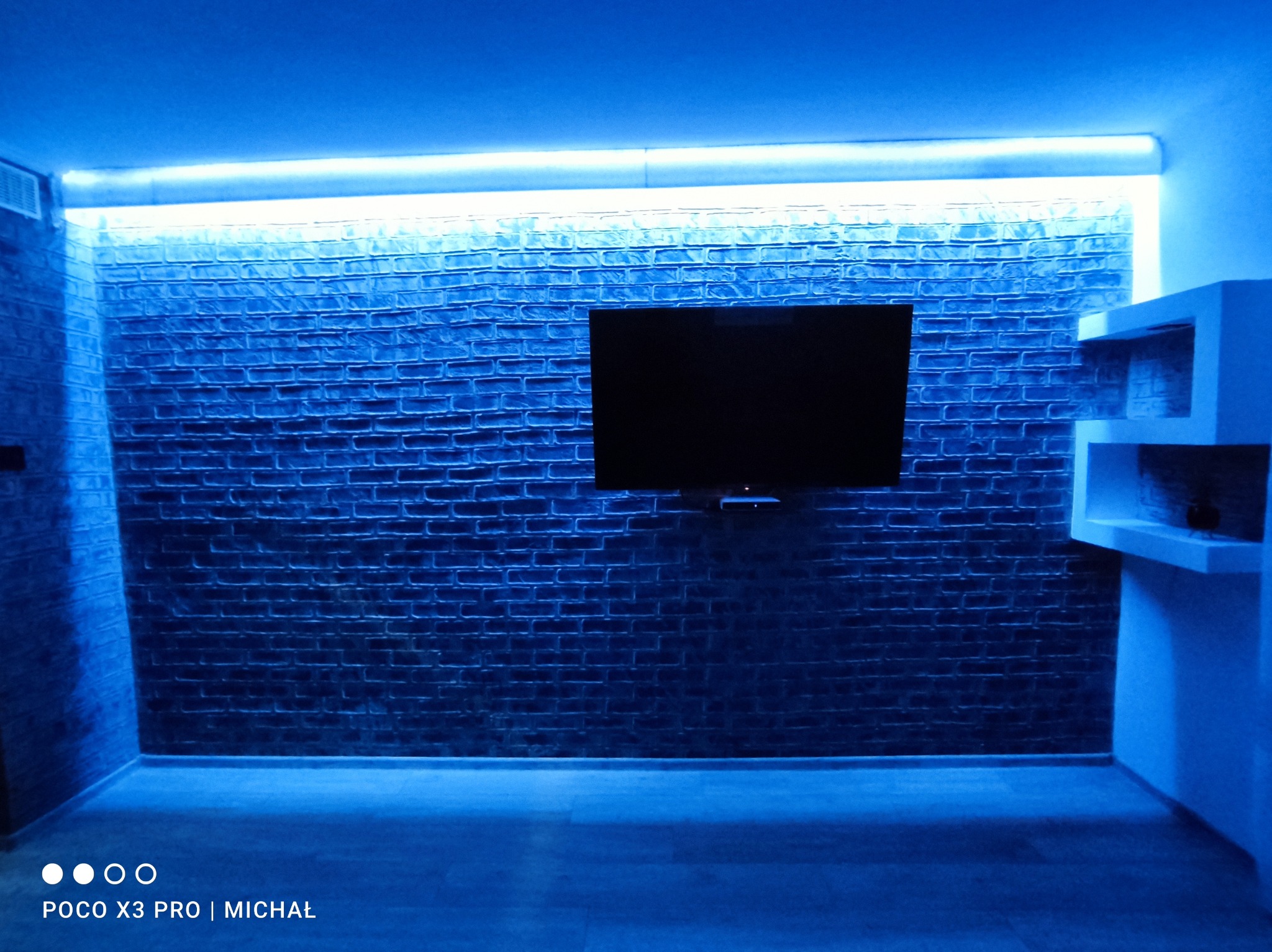 Pokój z ceglaną ścianą oświetloną niebieskim światłem LED, na ścianie wisi telewizor, obok półka. Nowoczesne wykończenie wnętrza z akcentem na oświetlenie.