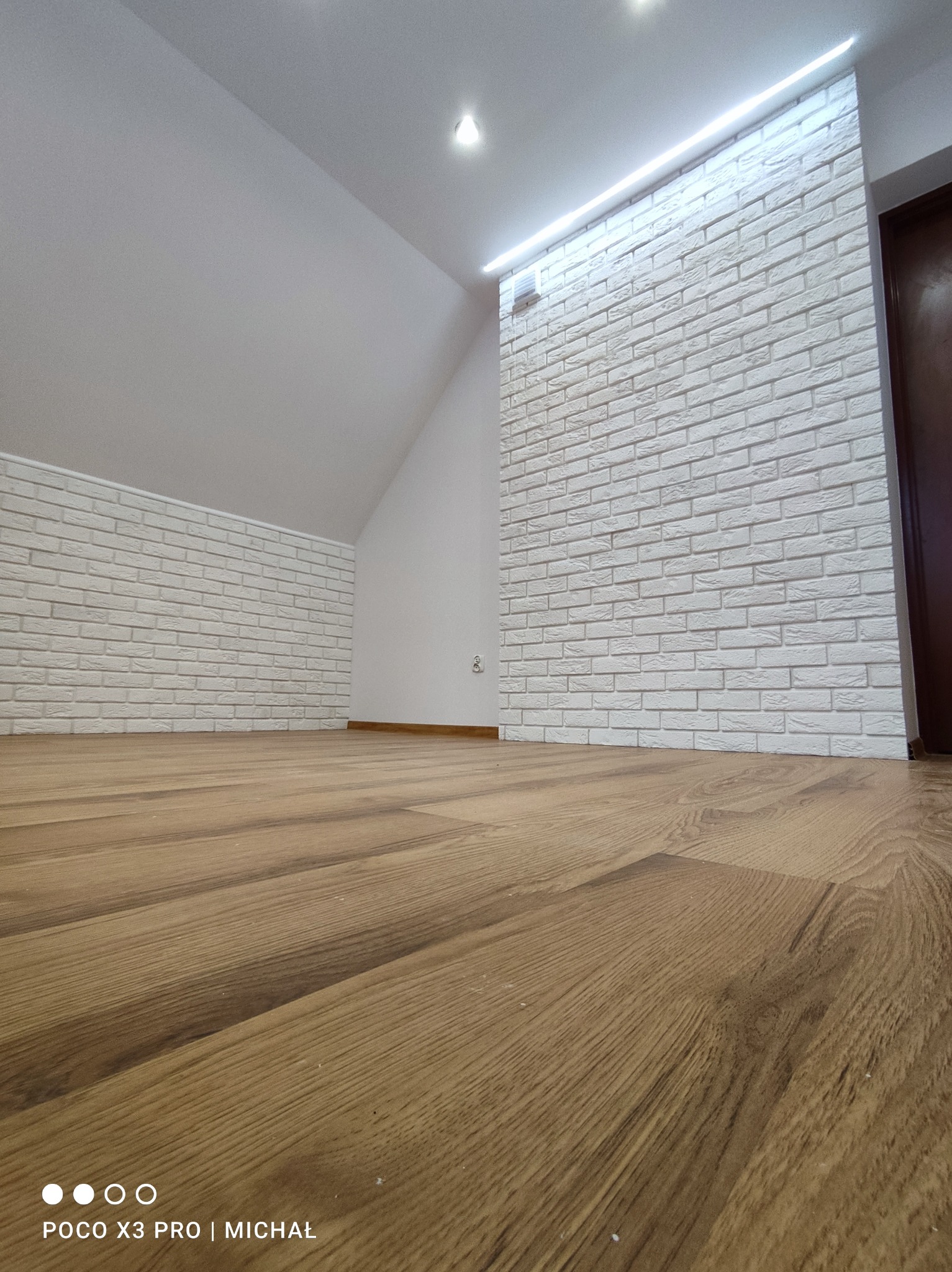 Poddasze po remoncie: drewniana podłoga, ściany wyłożone białą cegłą, oświetlenie LED przy suficie. Ujęcie z niskiej perspektywy podkreśla przestrzeń.