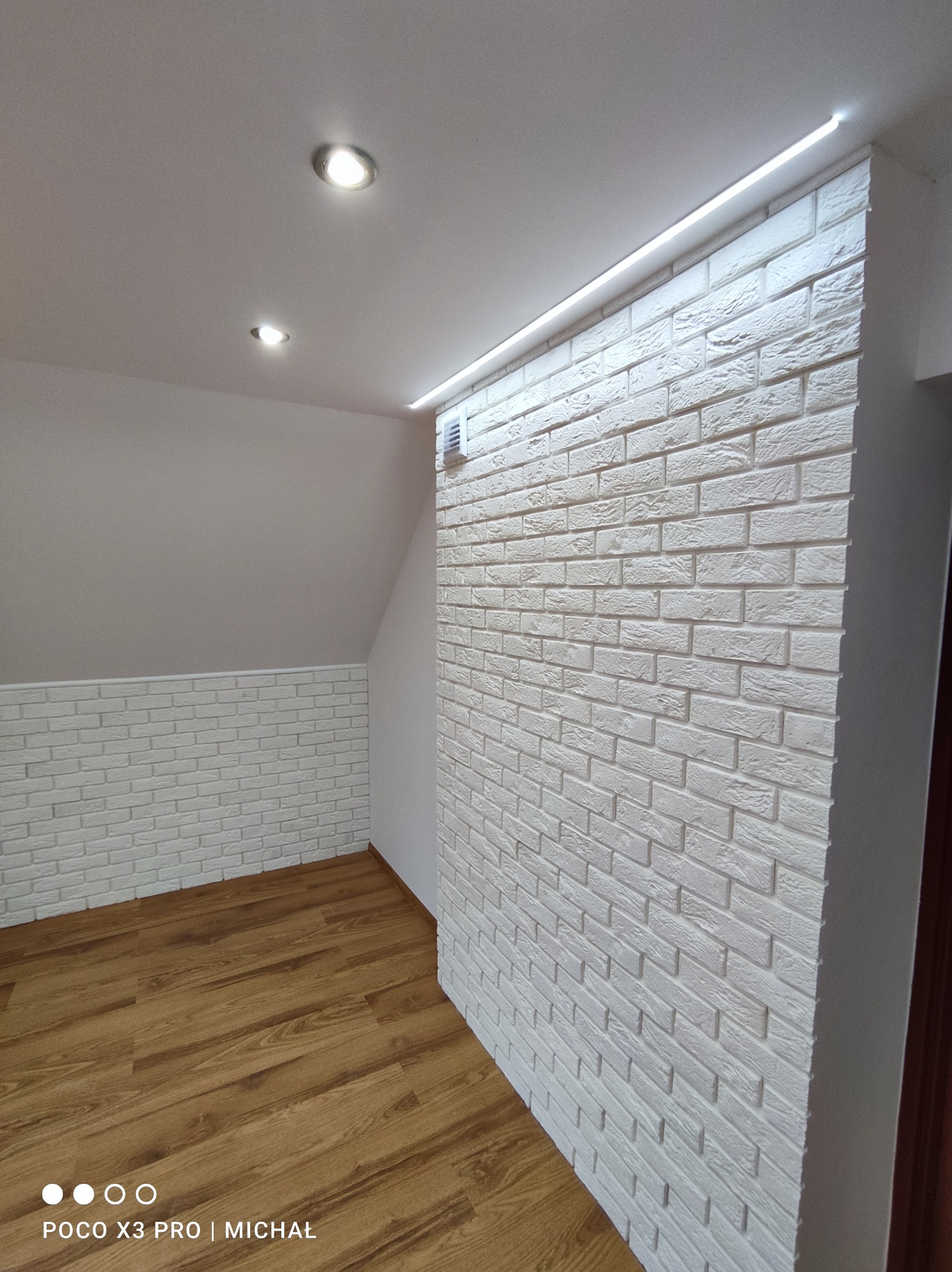 Wykończone poddasze z dekoracyjną białą cegłą na ścianie i podświetleniem LED, uzupełnione panelami podłogowymi w odcieniach brązu. Dwa punkty świetlne na suficie.