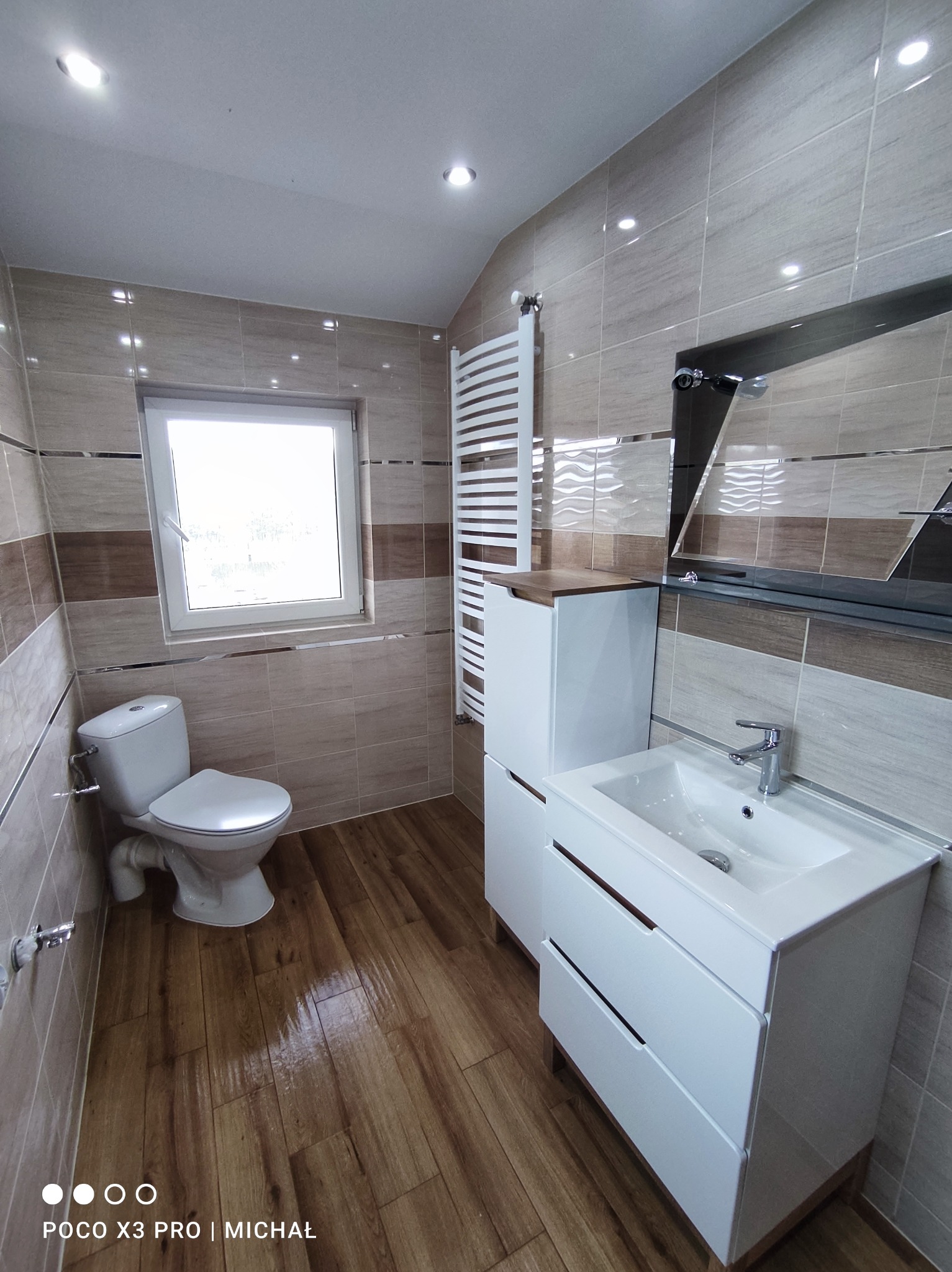 Jasna łazienka po remoncie: beżowe płytki z brązowym pasem, drewnopodobna podłoga, biała szafka z umywalką i toaleta. Grzejnik drabinkowy i lustro dopełniają wystrój.