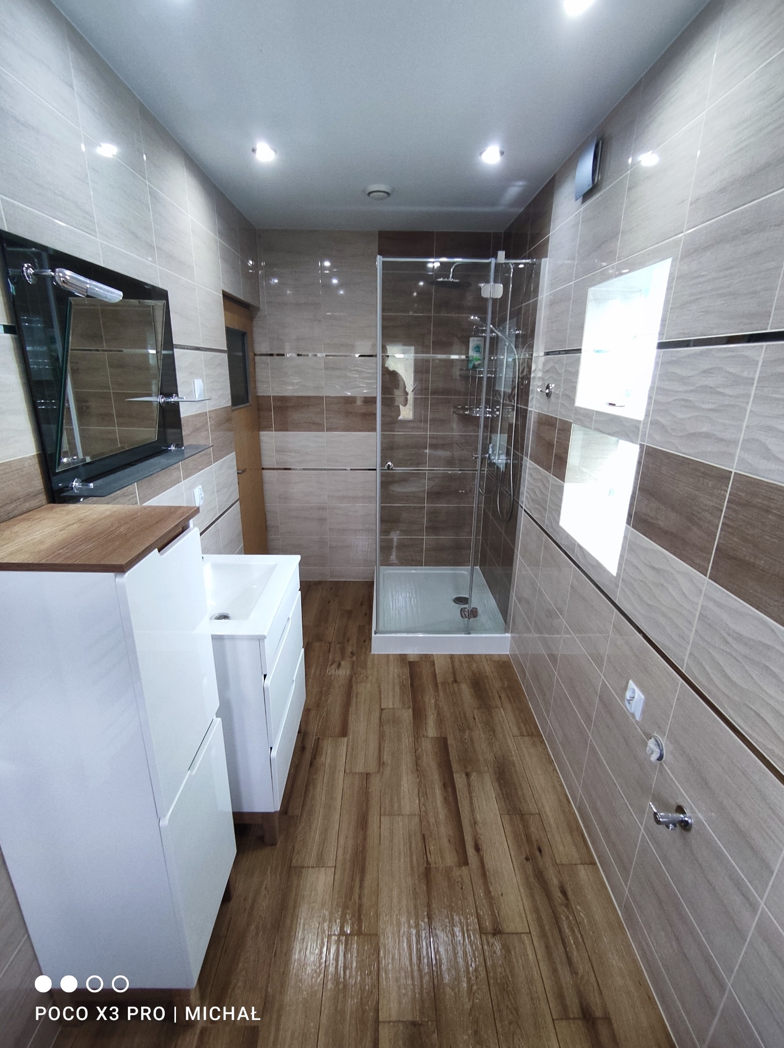 Wykończona łazienka z kabiną prysznicową, umywalką z szafką i lustrem. Na ścianach i podłodze płytki w odcieniach beżu i brązu. Widok na całe pomieszczenie.