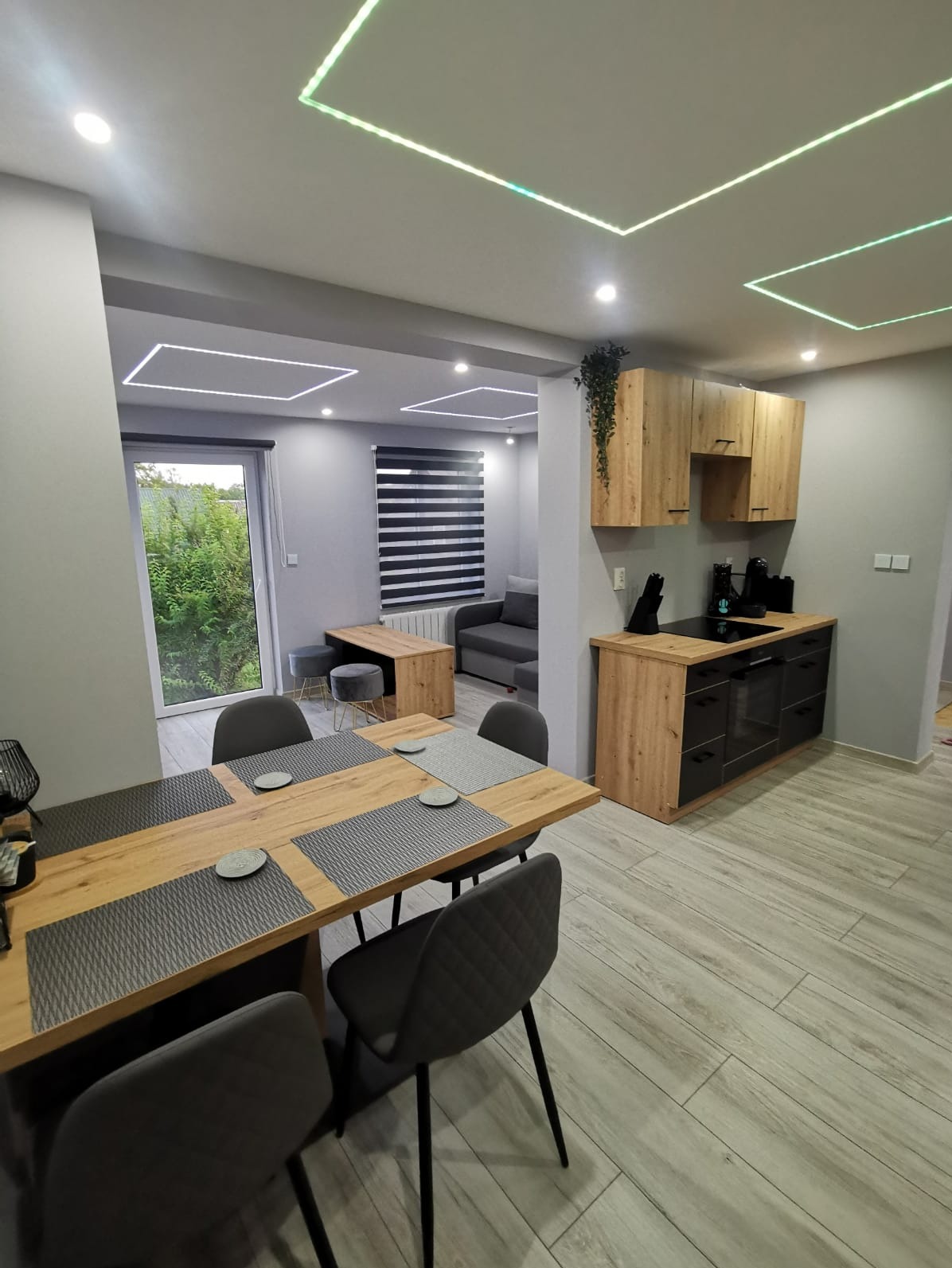 Nowoczesne wnętrze: drewniany stół z szarymi krzesłami, aneks kuchenny z drewnianymi szafkami i czarnymi frontami, podświetlany sufit, szare ściany i drewnopodobna podłoga.