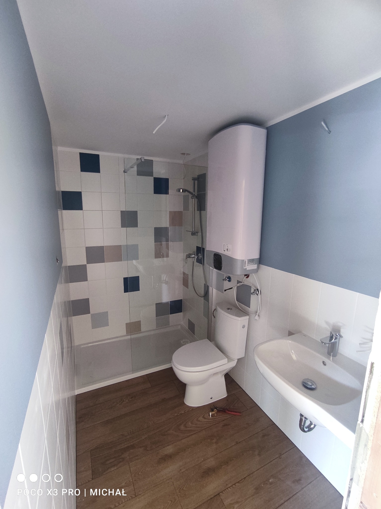 Wykończona łazienka z mozaiką nad wanną, drewnopodobną podłogą i białymi sanitariatami. Widoczny bojler, umywalka i toaleta. Częściowo widoczne narzędzia na podłodze.