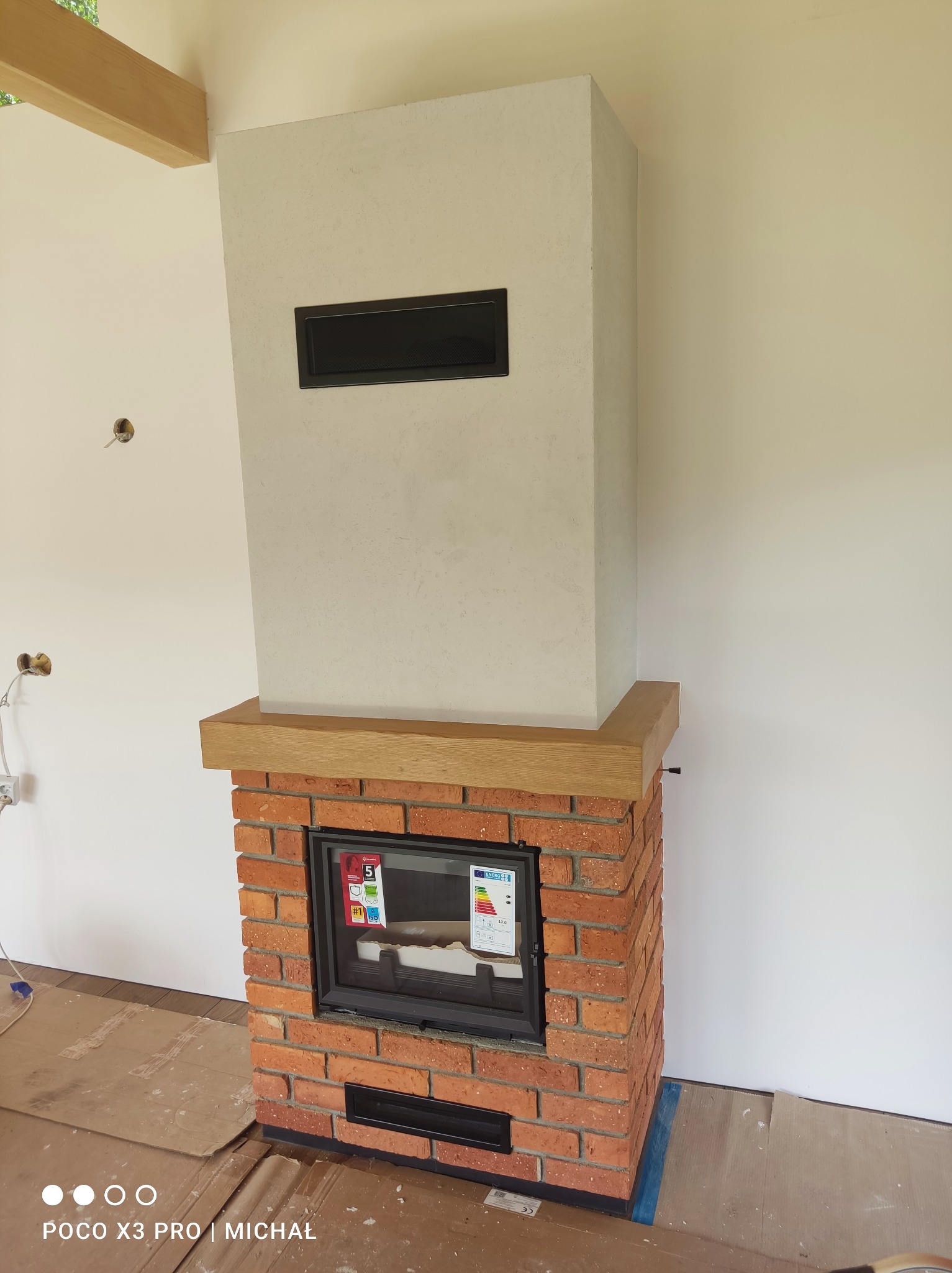 Murowany kominek z cegły i elementami drewna, z wkładem kominkowym, na tle białej ściany. Widoczne zabezpieczenia podłogi. Detal wykończenia wnętrza.